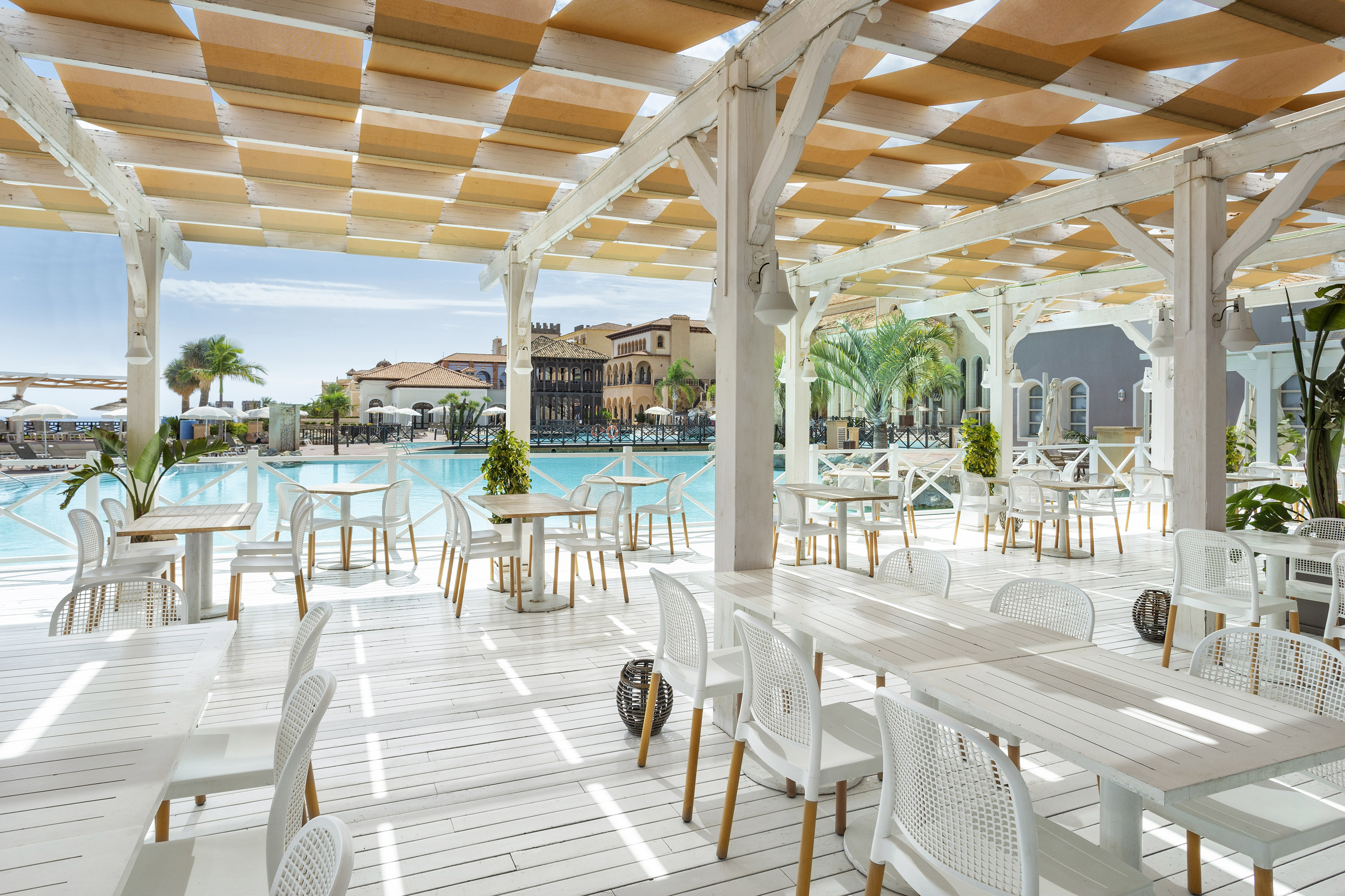 Resort outdoor dining patio with white furniture, pool, and shaded canopy.
