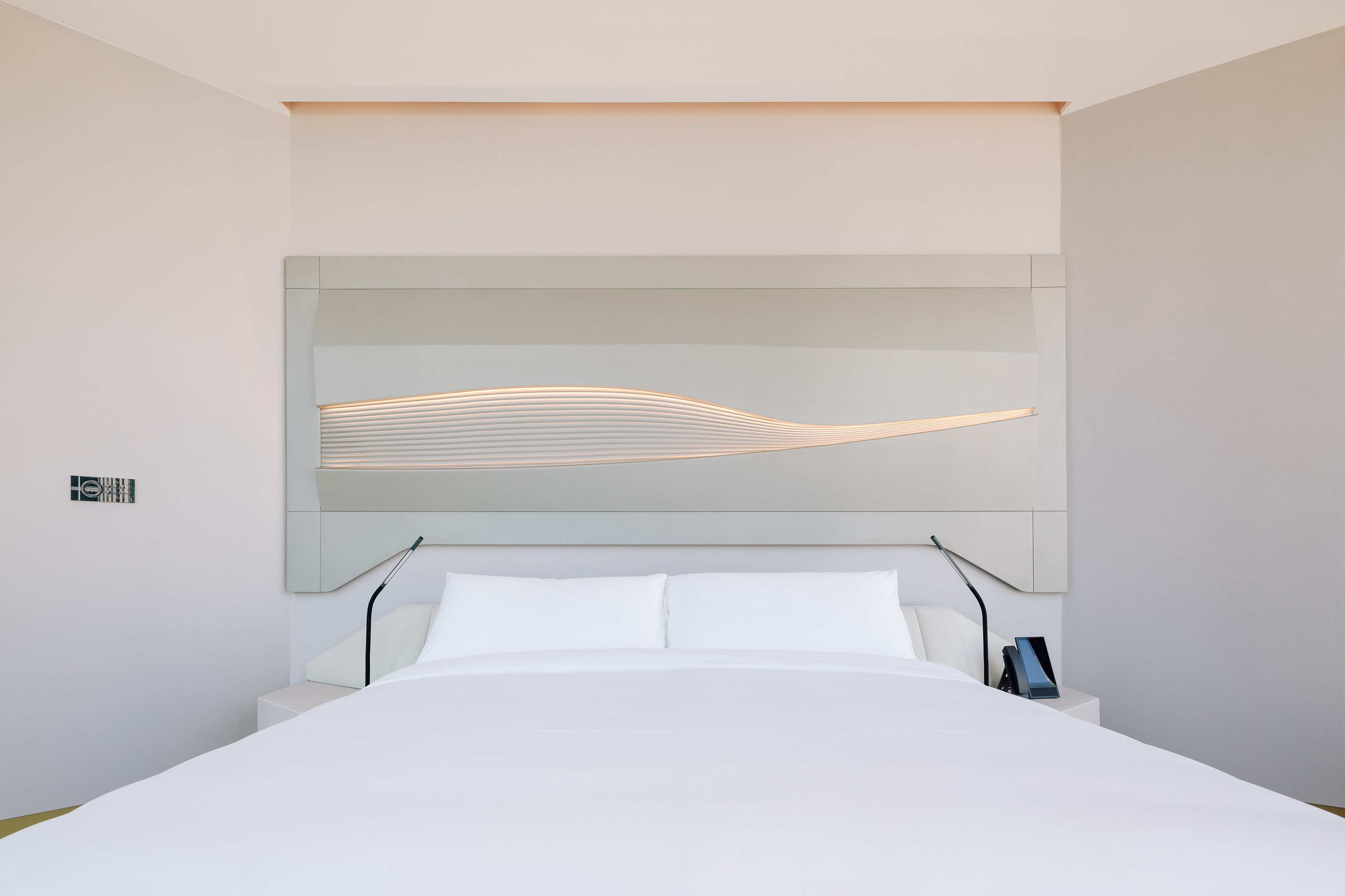 Modern hotel bedroom with white bed and illuminated wavy headboard.