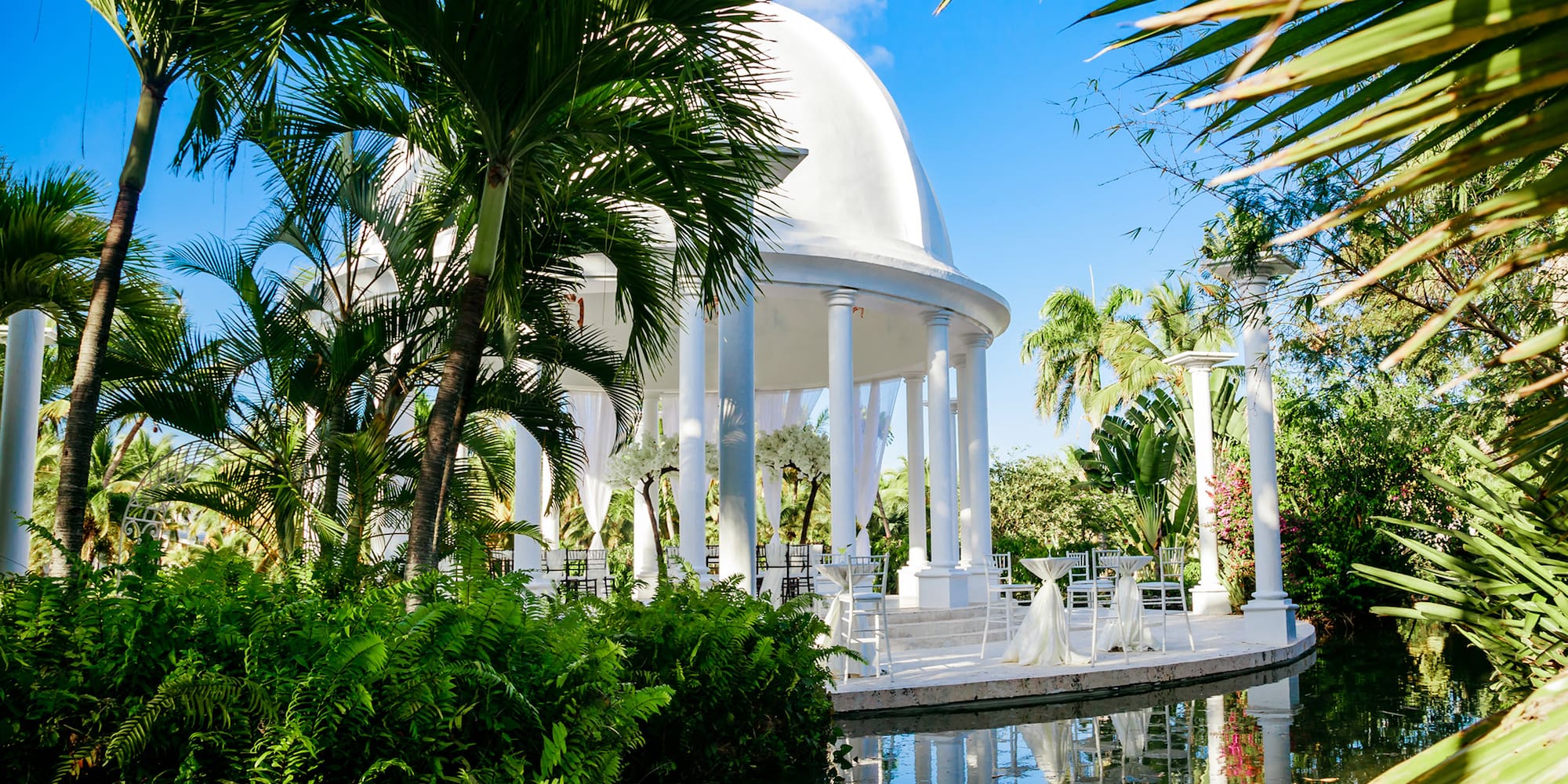 a white gazebo with white columns and a white dome with chairs and a pool of water