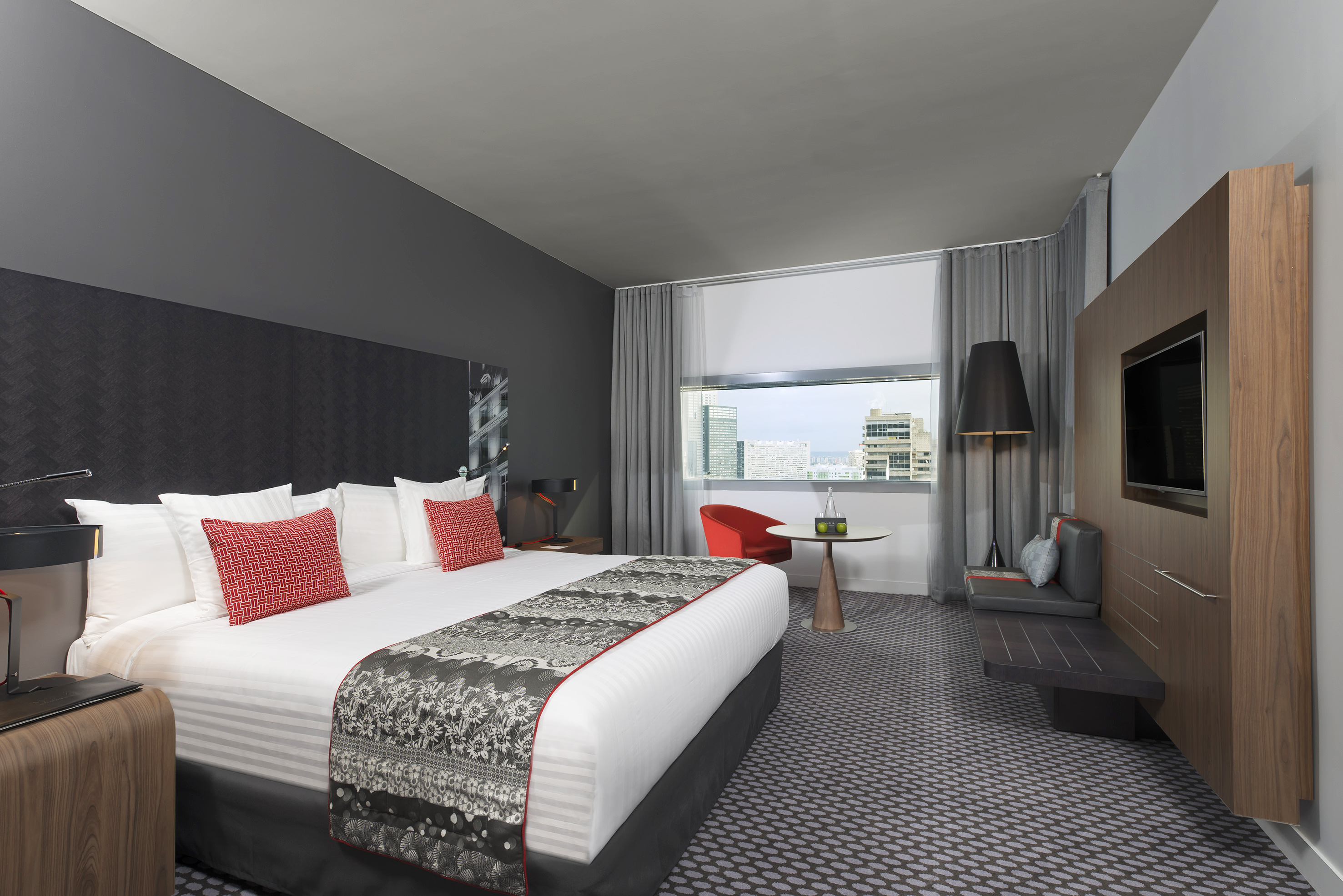 Modern hotel bedroom with king bed, city view, and red accents.