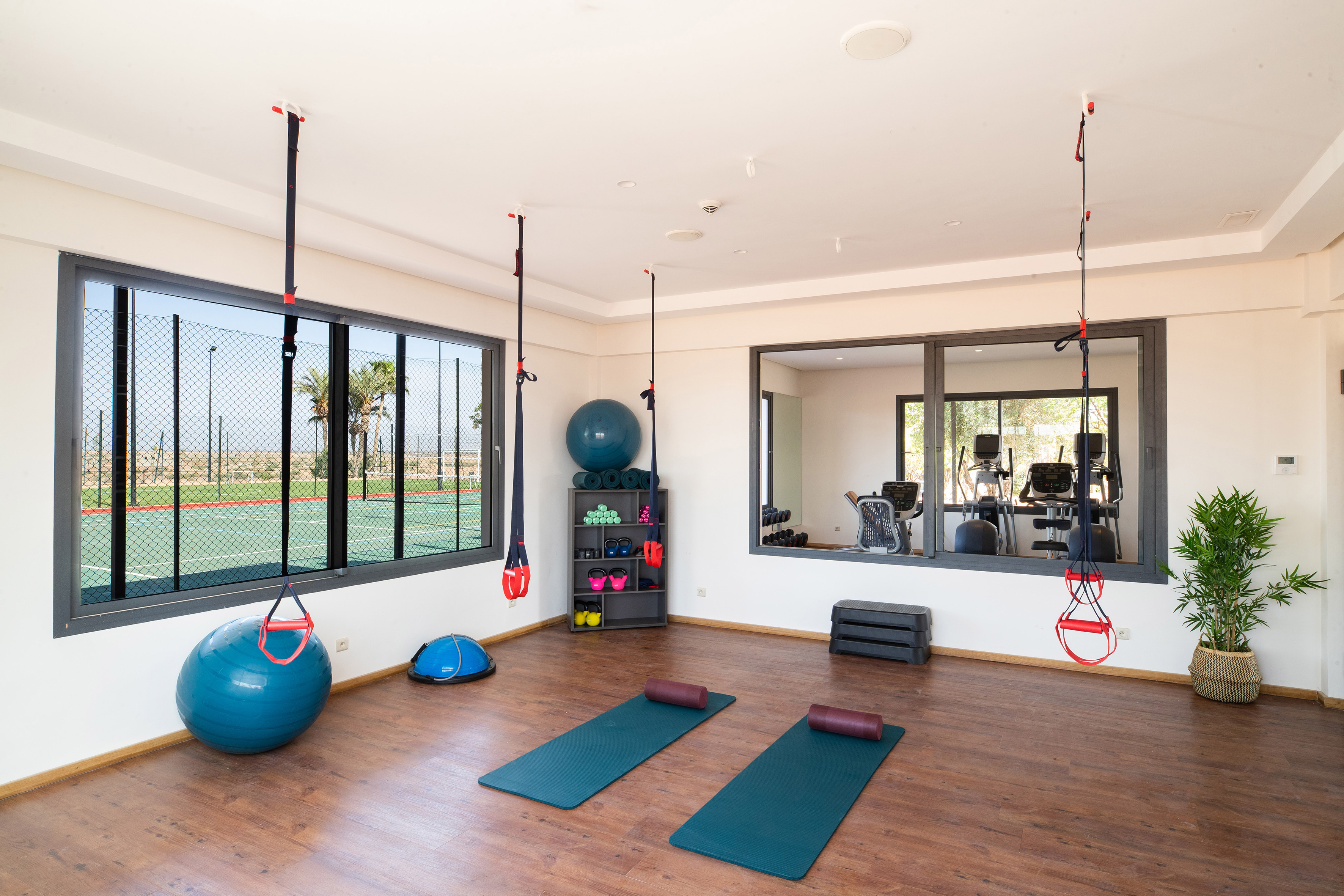 Fitness room with exercise equipment, yoga mats, and a tennis court view.