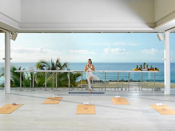 a woman standing on one leg in yoga pose on mat on a balcony overlooking the ocean