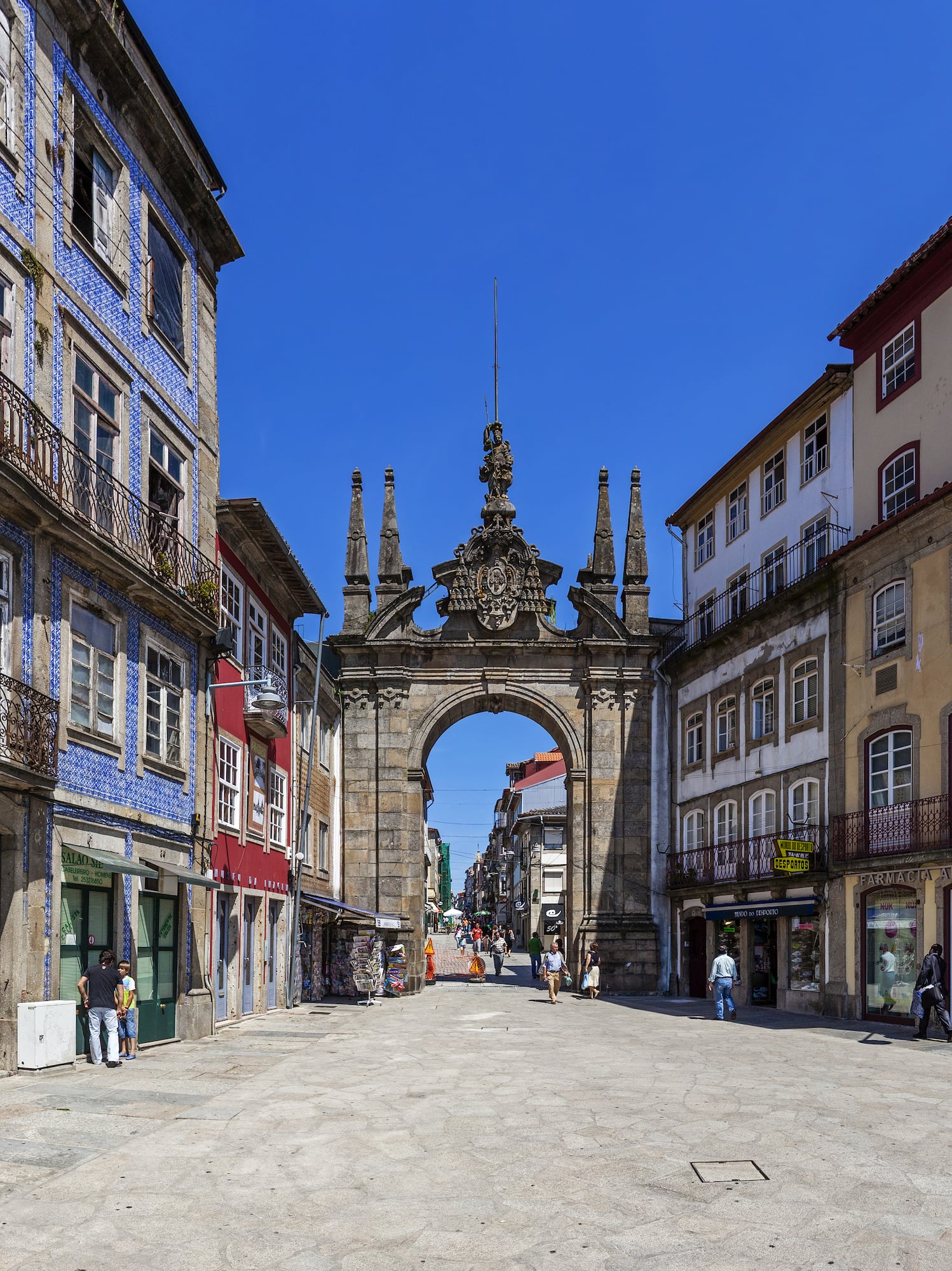a stone archway in a street with buildings and people walking with Arco da Porta Nova in the background
