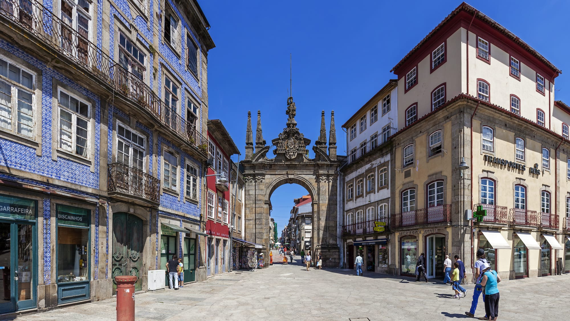 a stone archway in a street with buildings and people walking with Arco da Porta Nova in the background
