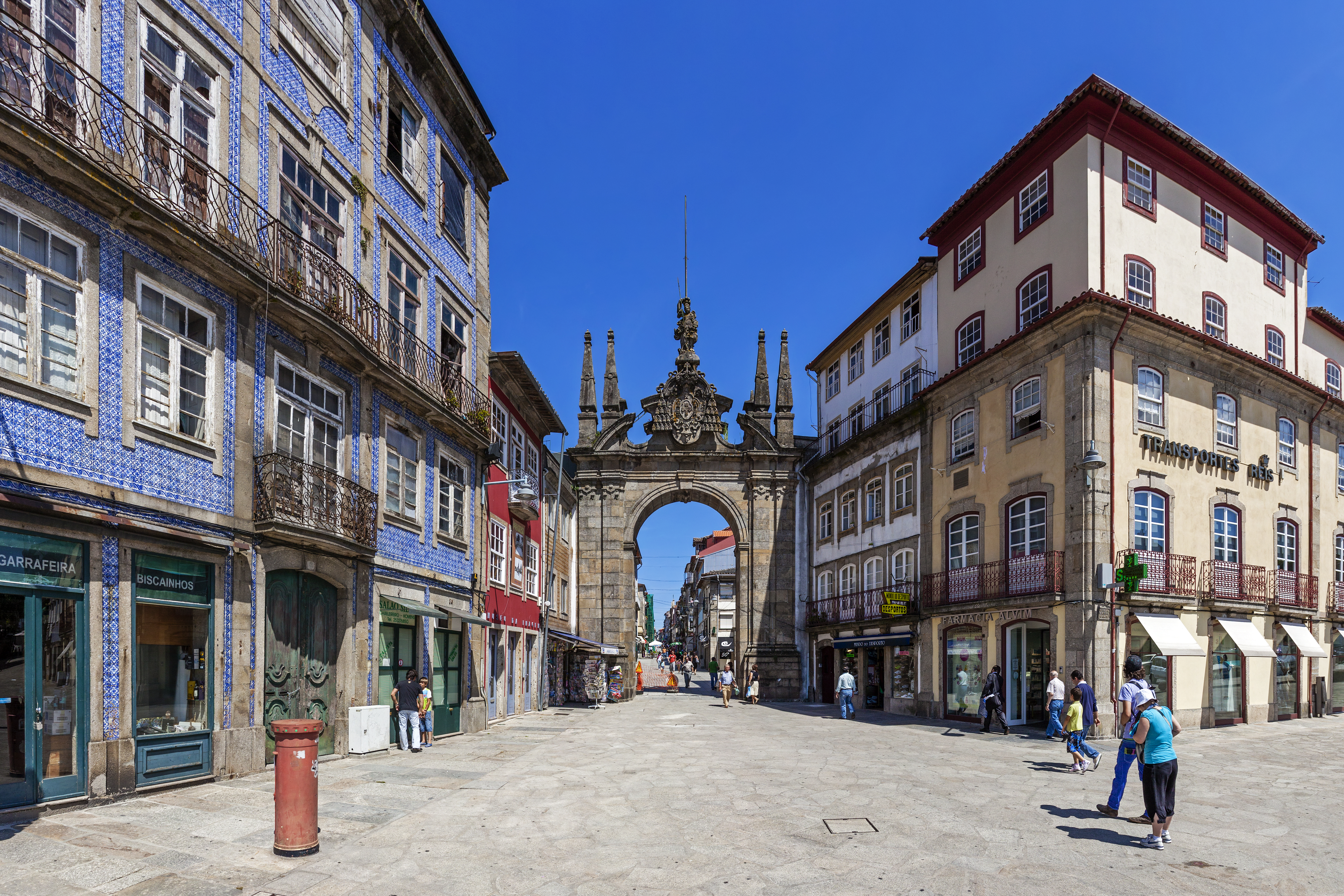a stone archway in a street with buildings and people walking with Arco da Porta Nova in the background