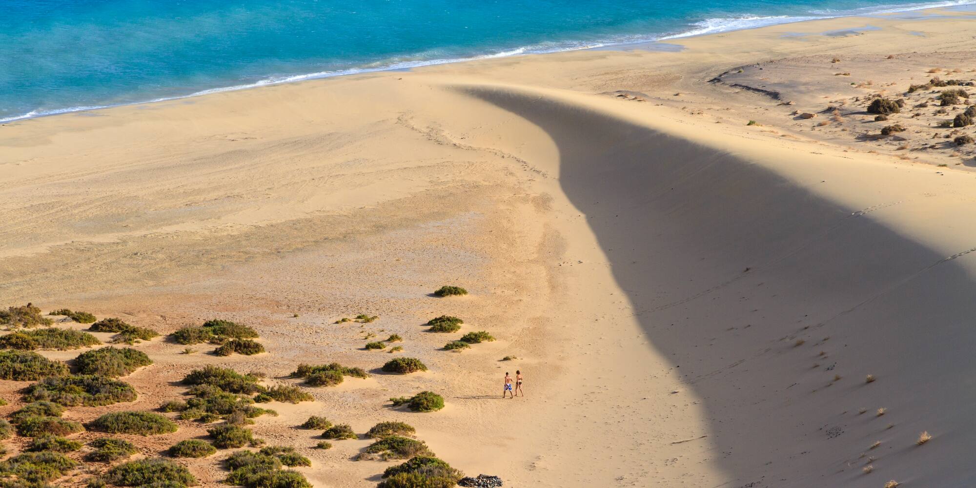 Vast sandy beach with blue ocean, large dune, and two people walking.