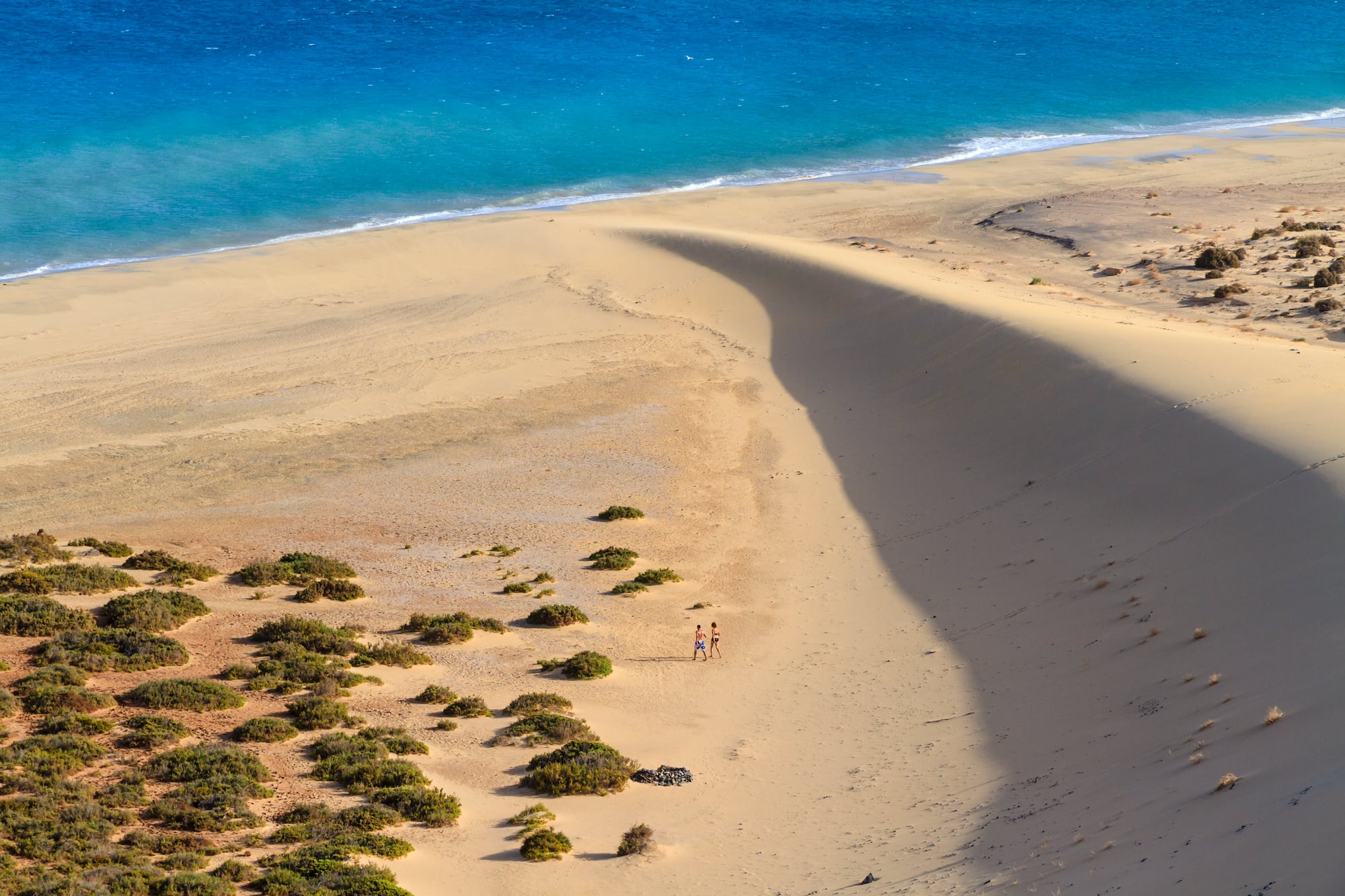 Playa de arena con dunas, océano azul y dos personas caminando.