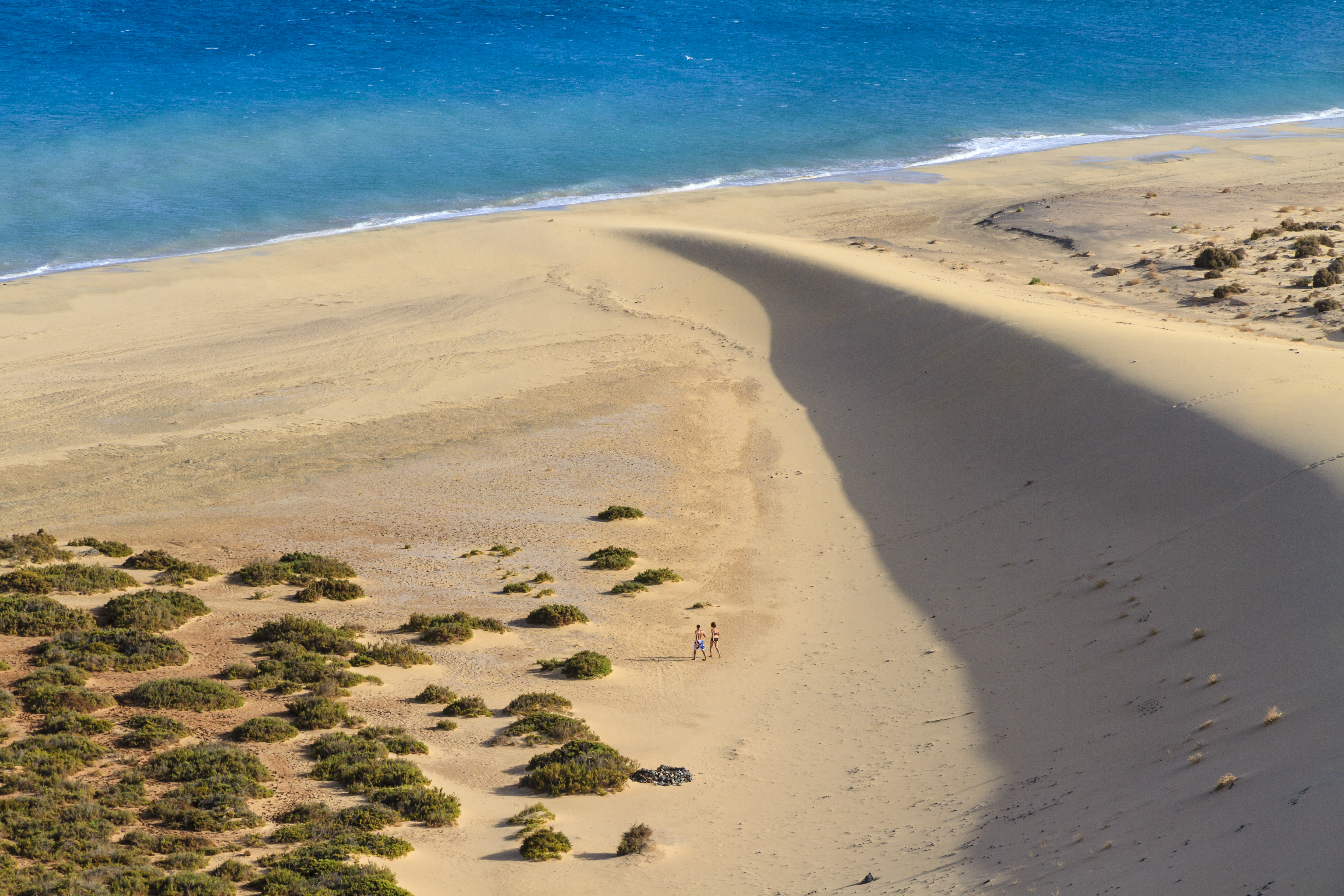 Vast sandy beach with blue ocean, large dune, and two people walking.