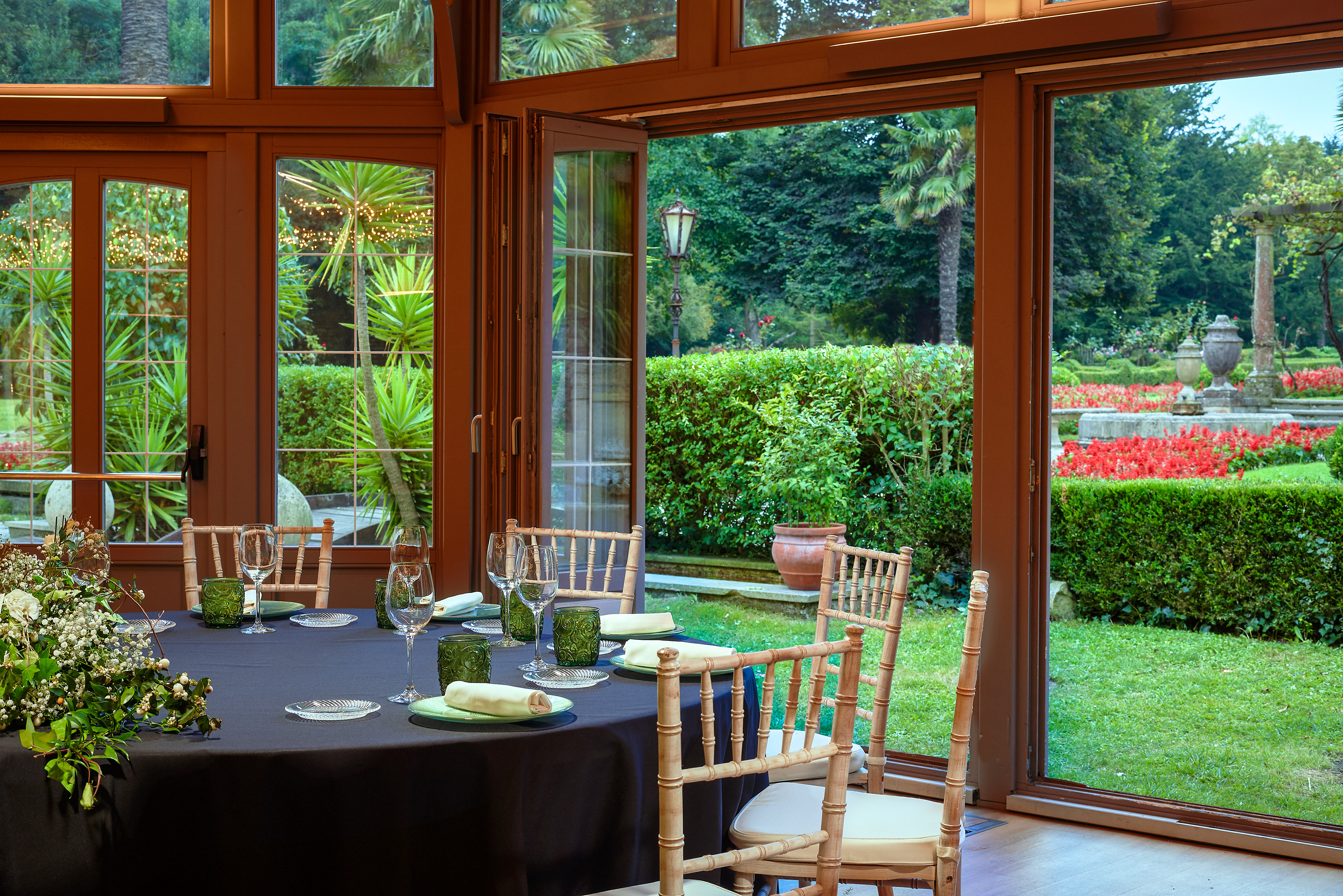 Elegant dining table set in glass room overlooking lush garden with red flowers. - Salón del Jardín - Palacio de Avilés Afiliat Meliá, Avilés