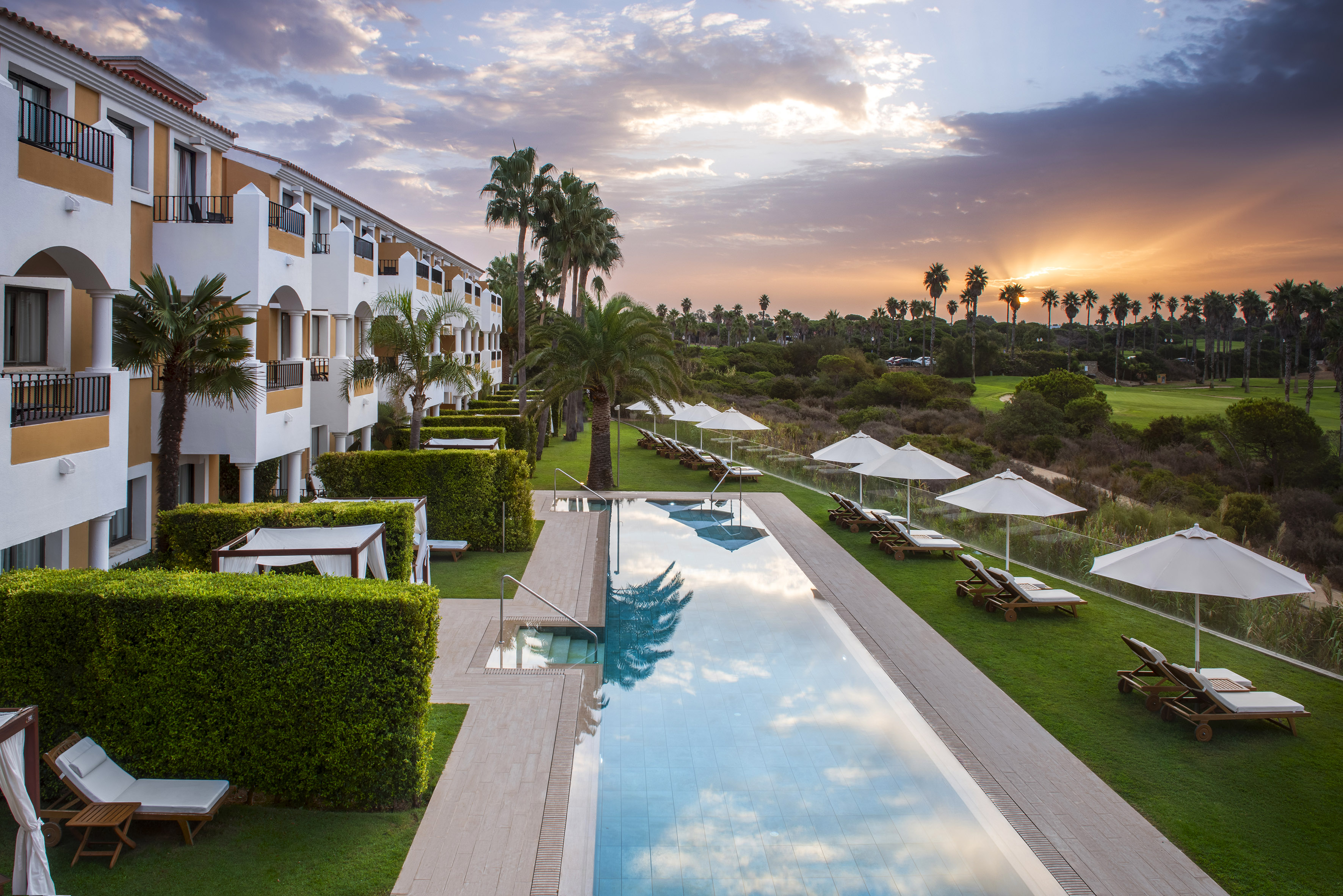 Luxury resort hotel with swimming pool, palm trees, and golf course at sunset.