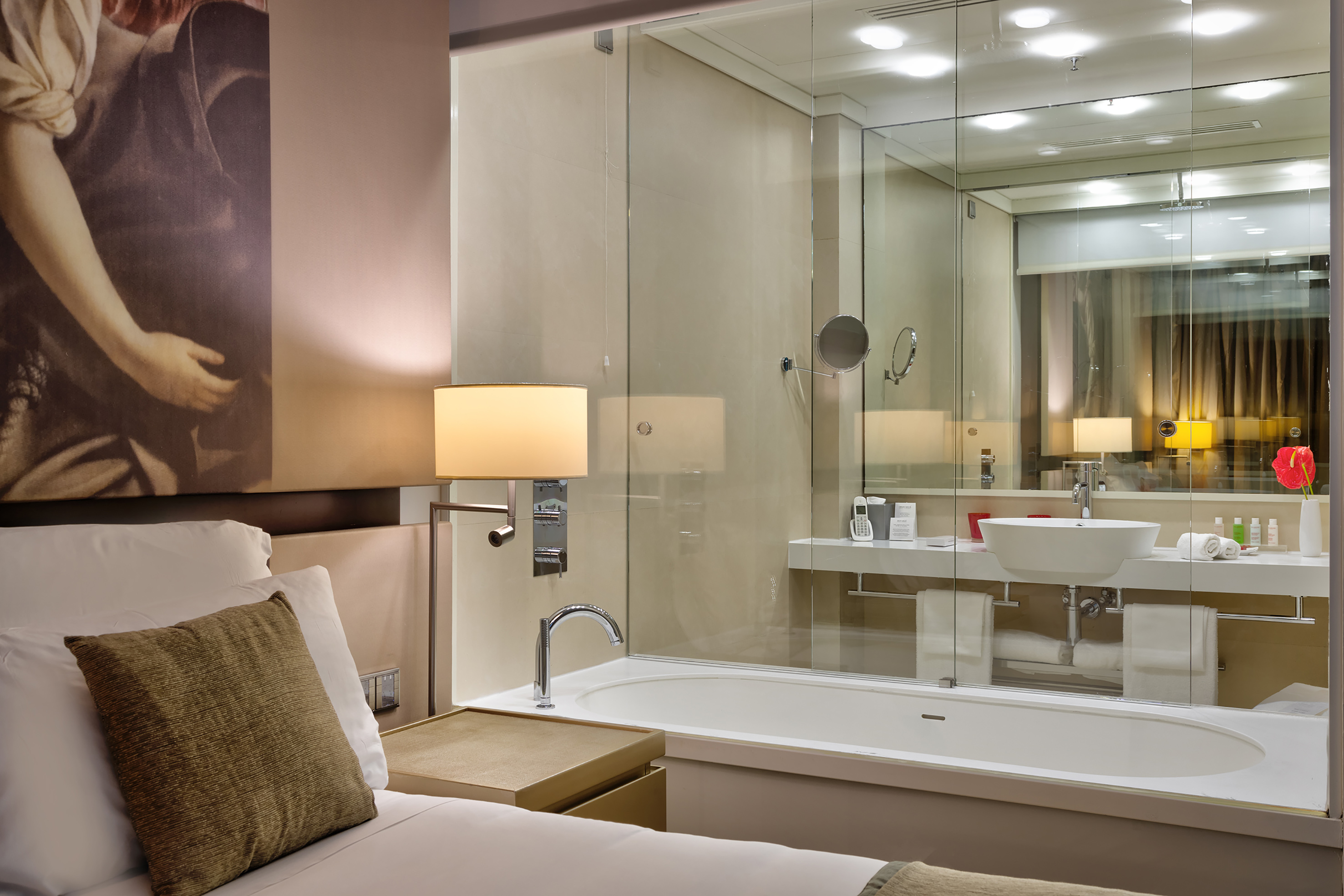 Modern hotel bedroom with glass-walled bathroom, bathtub, and illuminated lamp.