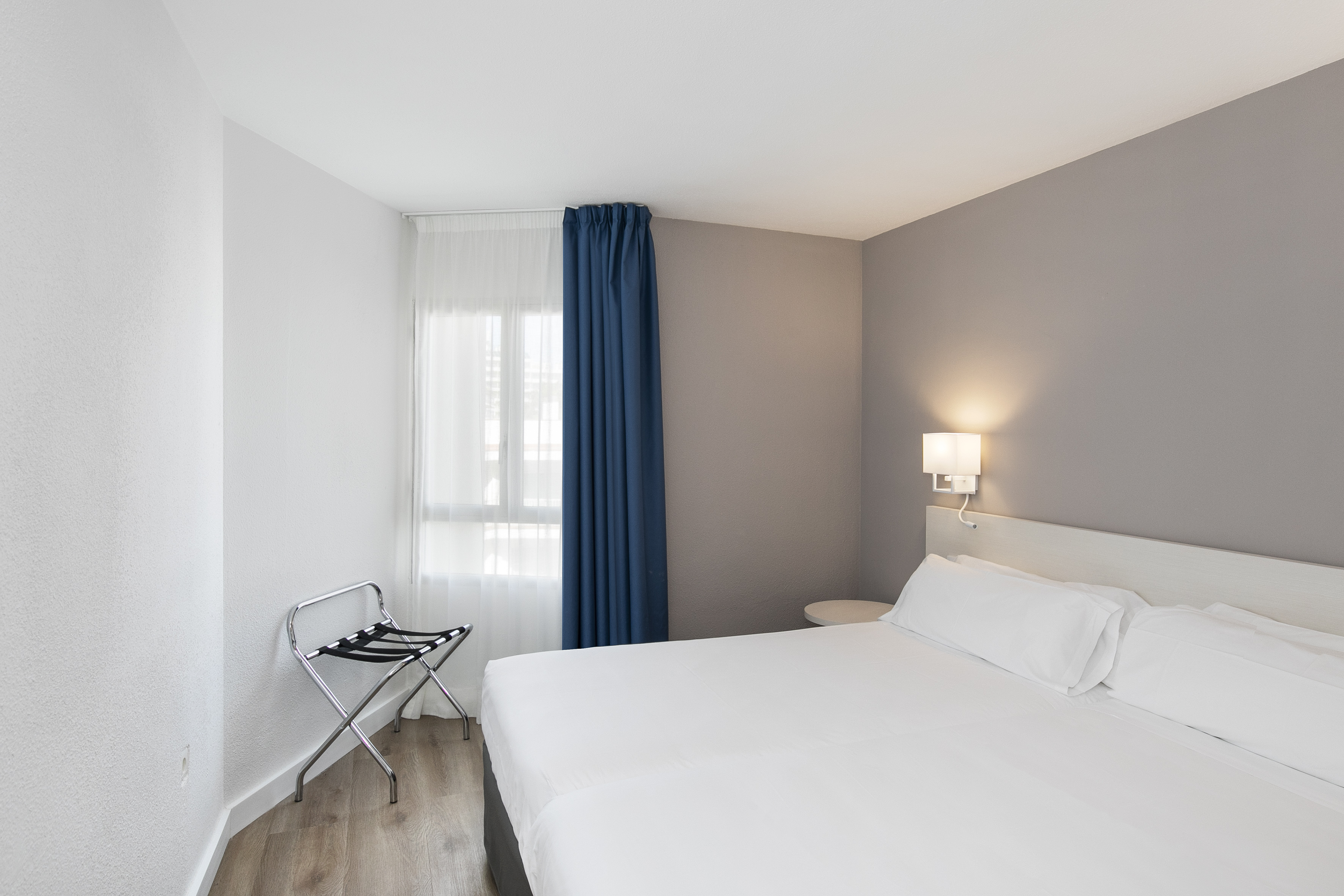 Modern hotel bedroom with white bed, blue curtains, and luggage rack.