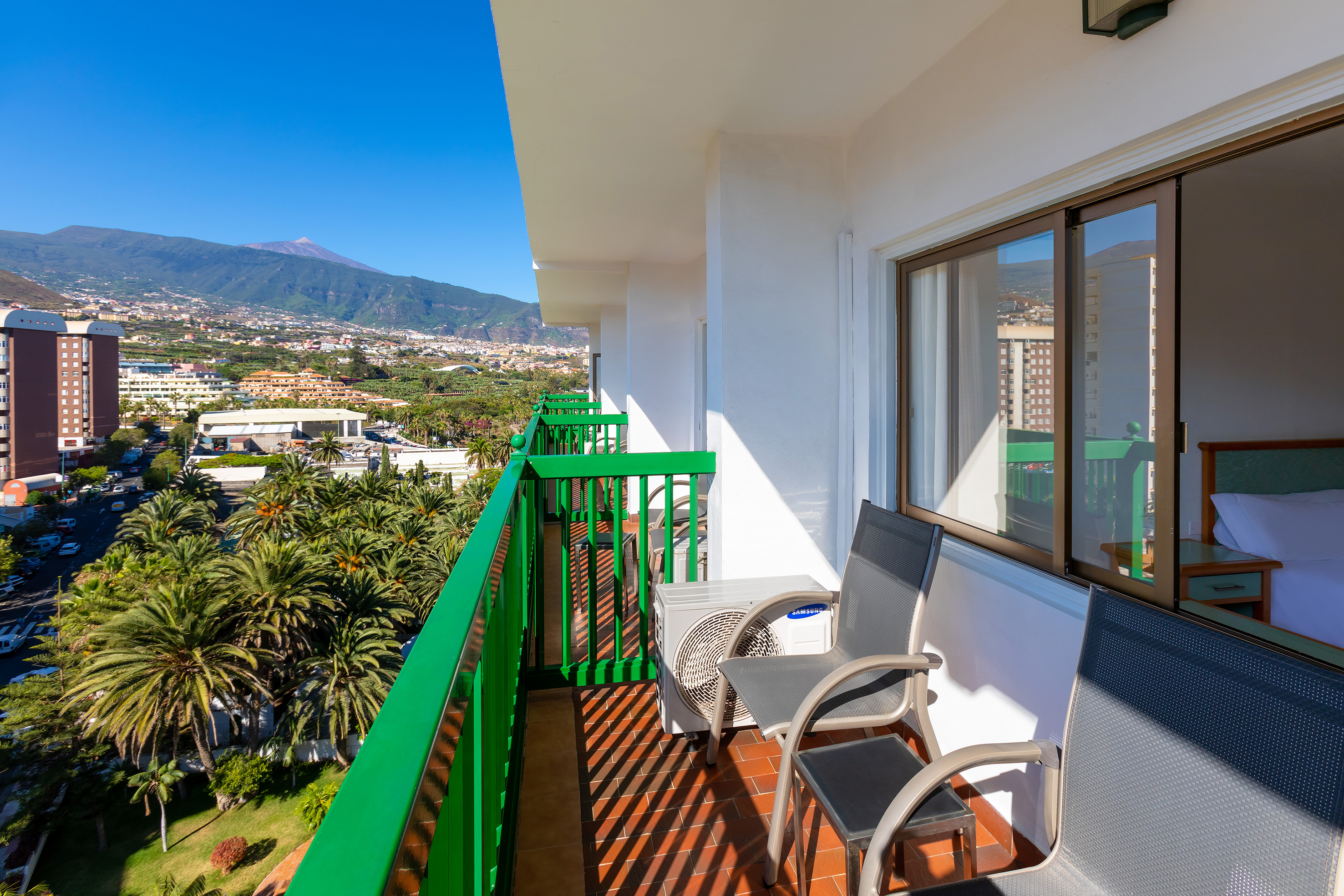 Sunny hotel balcony with chairs, palm trees, city, and distant mountain views.