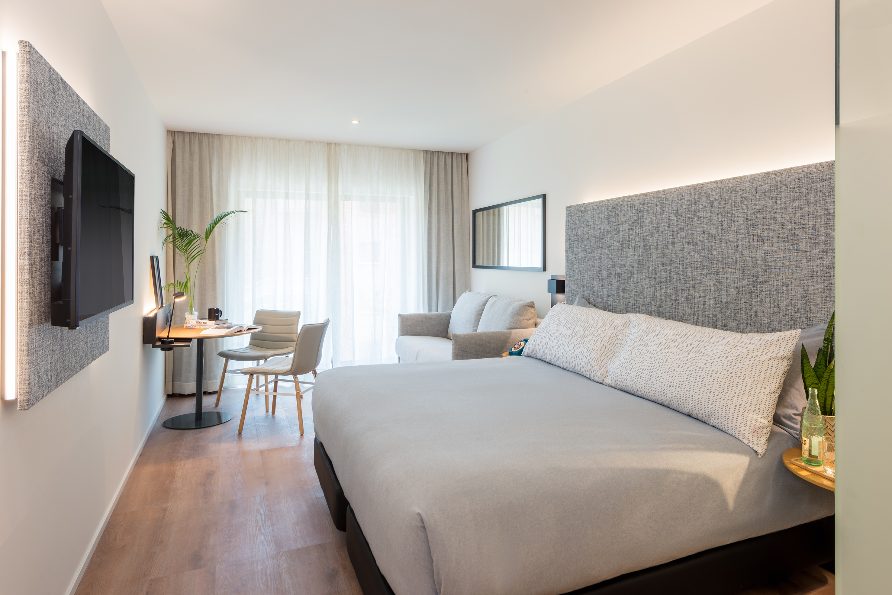 Modern hotel room with king bed, TV, dining area, sofa, and bright windows.