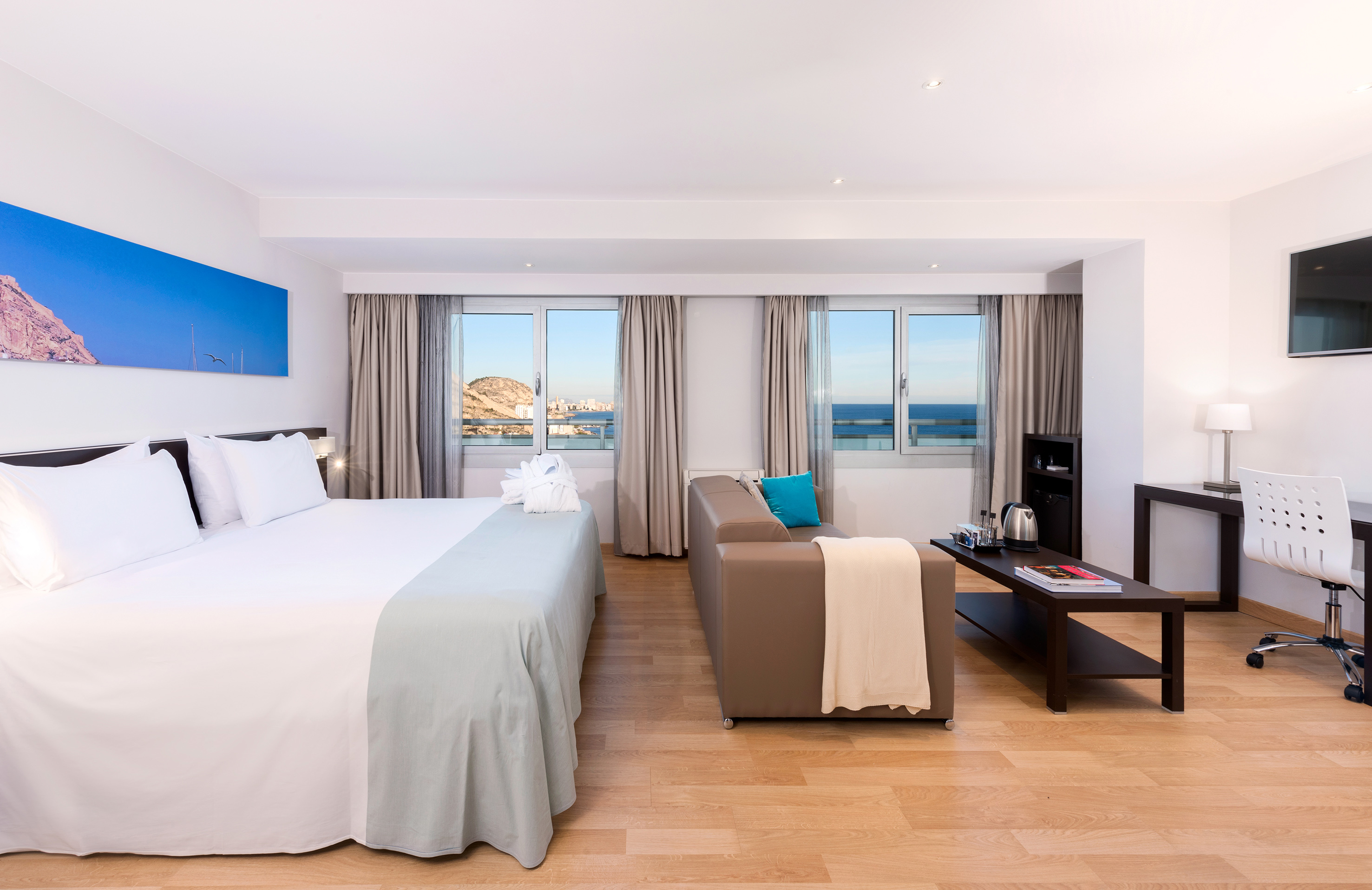 Modern hotel room with sea view, large bed, sofa, and desk by bright windows.