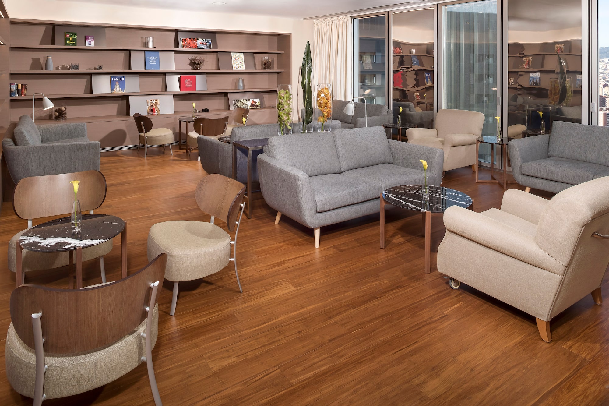 Hotel lounge with modern gray sofas, beige armchairs, wood floor, and bookshelves.
