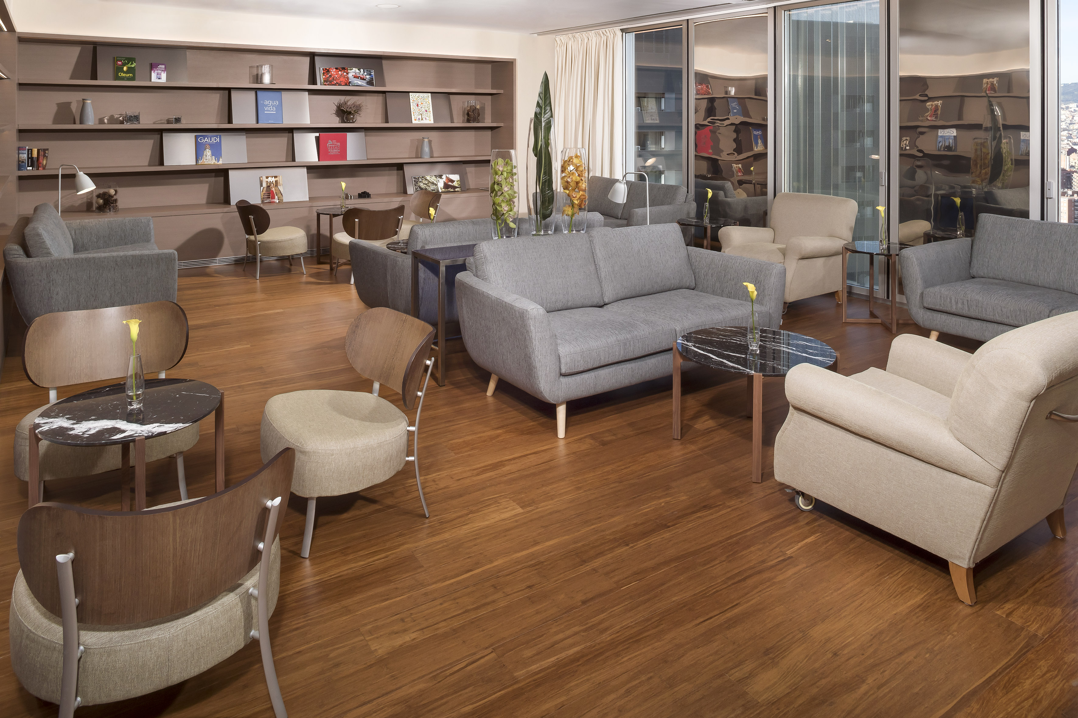 Hotel lounge with modern gray sofas, beige armchairs, wood floor, and bookshelves.