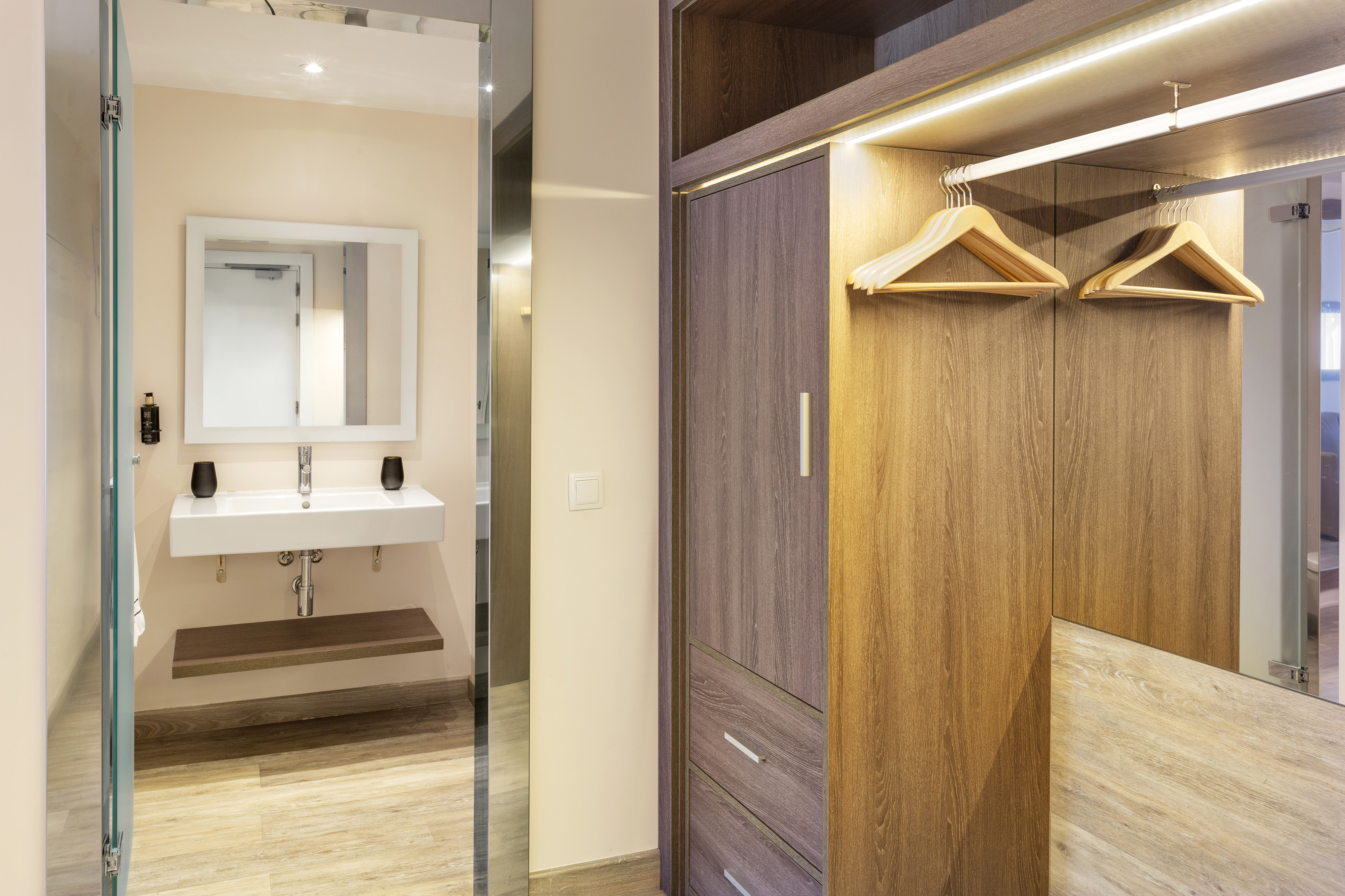Modern hotel room with integrated bathroom vanity and illuminated wood wardrobe.