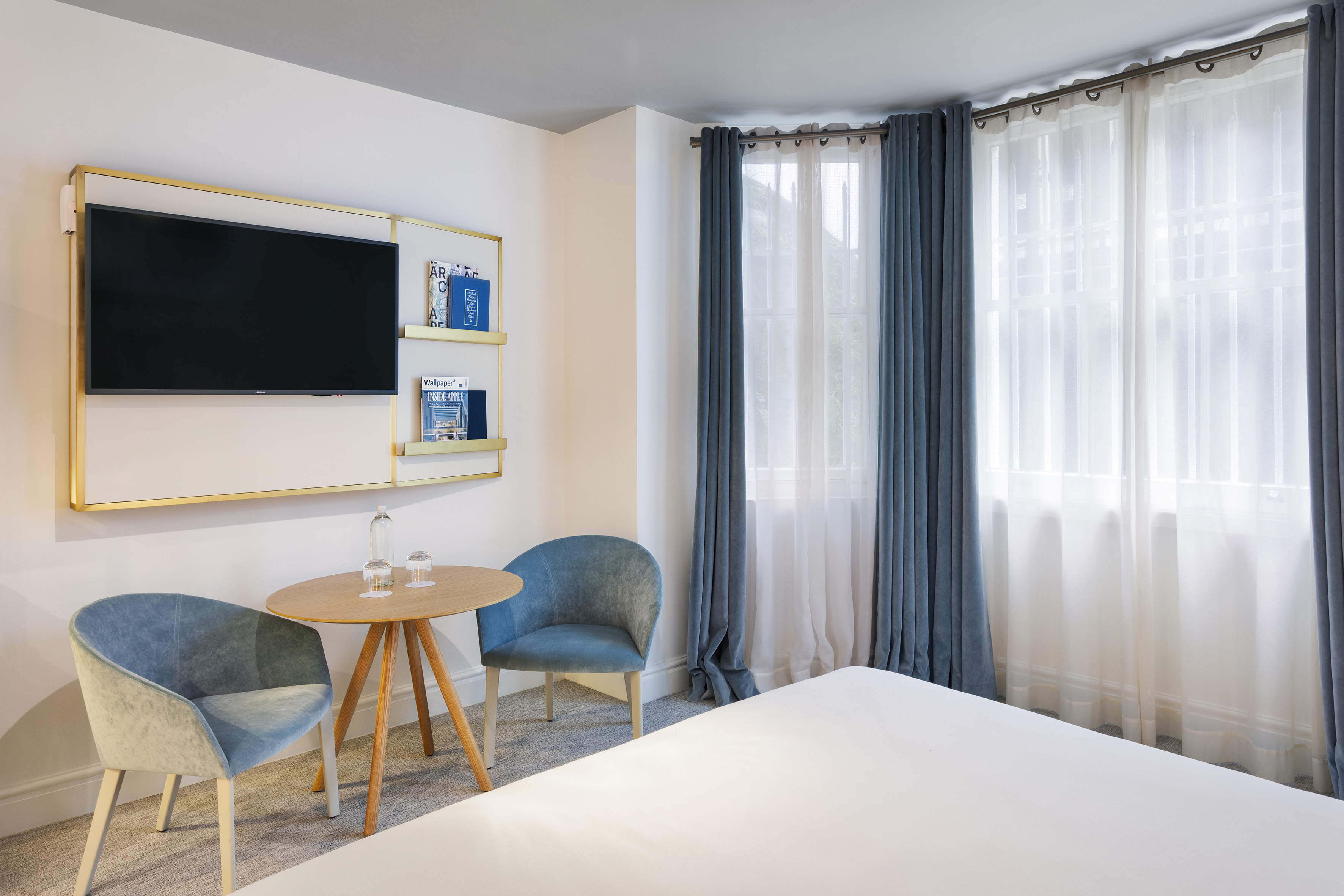 Modern hotel bedroom with blue chairs, round table, wall TV, and window.