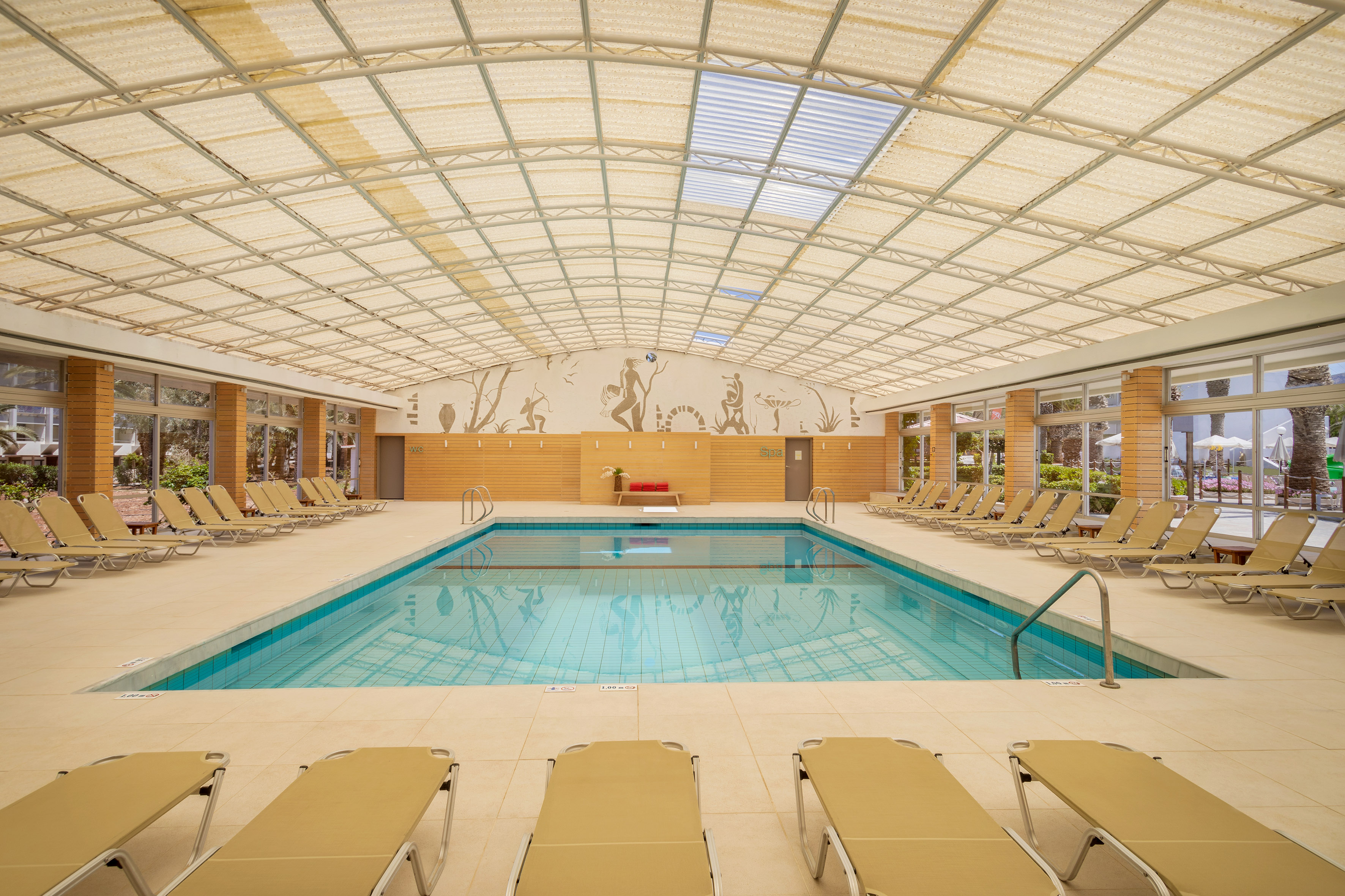 Covered hotel swimming pool with blue water, sun loungers, and large windows.