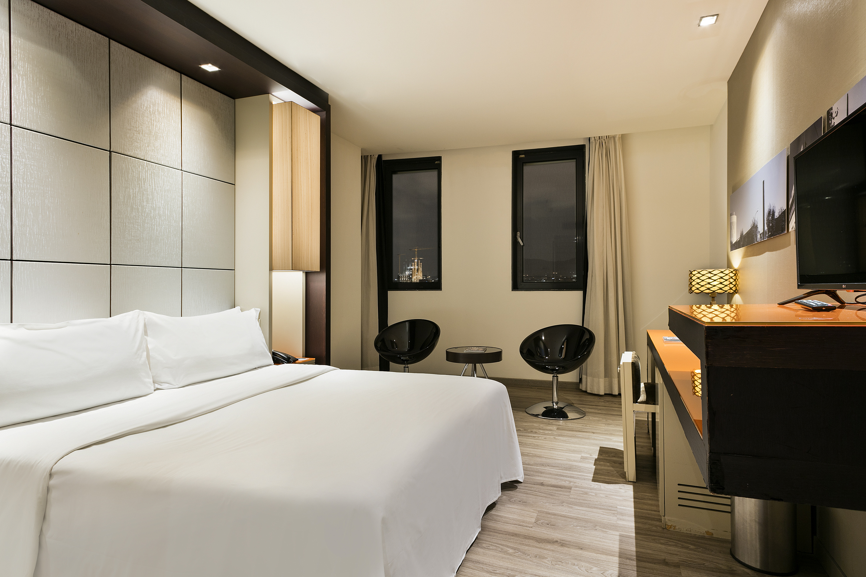 Contemporary hotel bedroom, white bed, city view, and sleek black chairs.