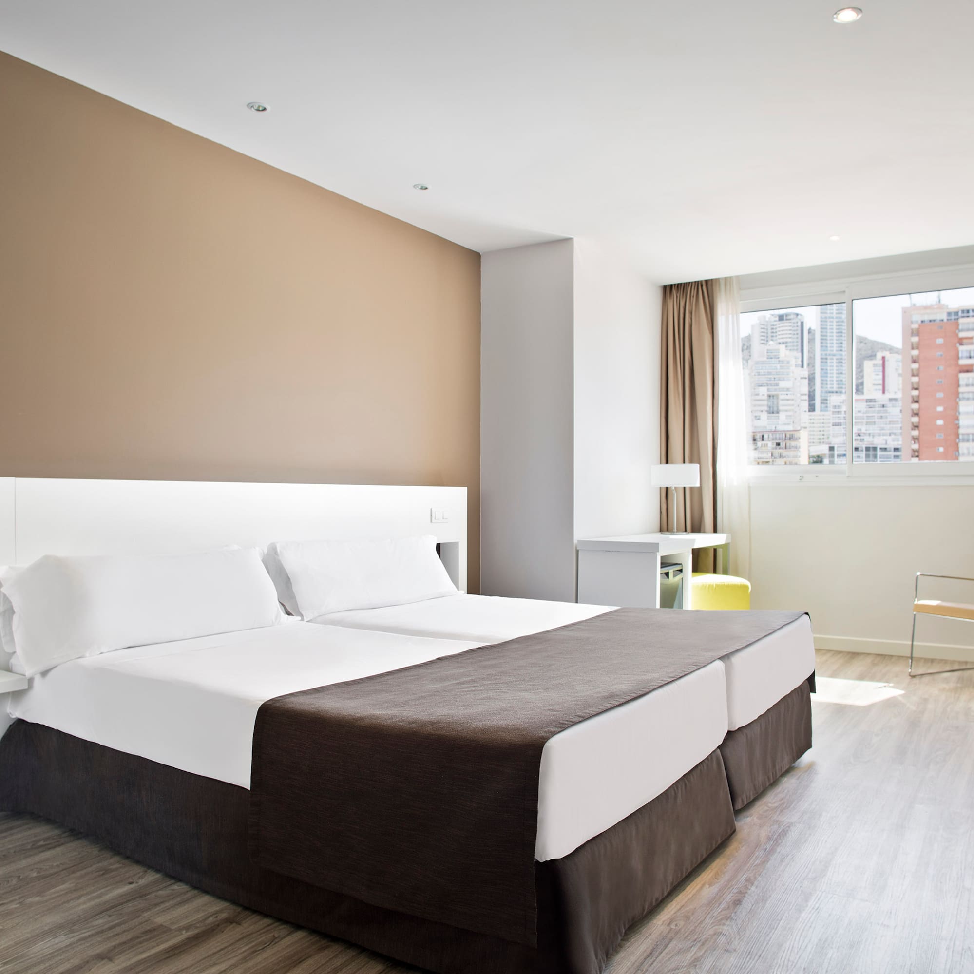 Modern hotel room with twin beds, city view, light wood floor, and bright decor.