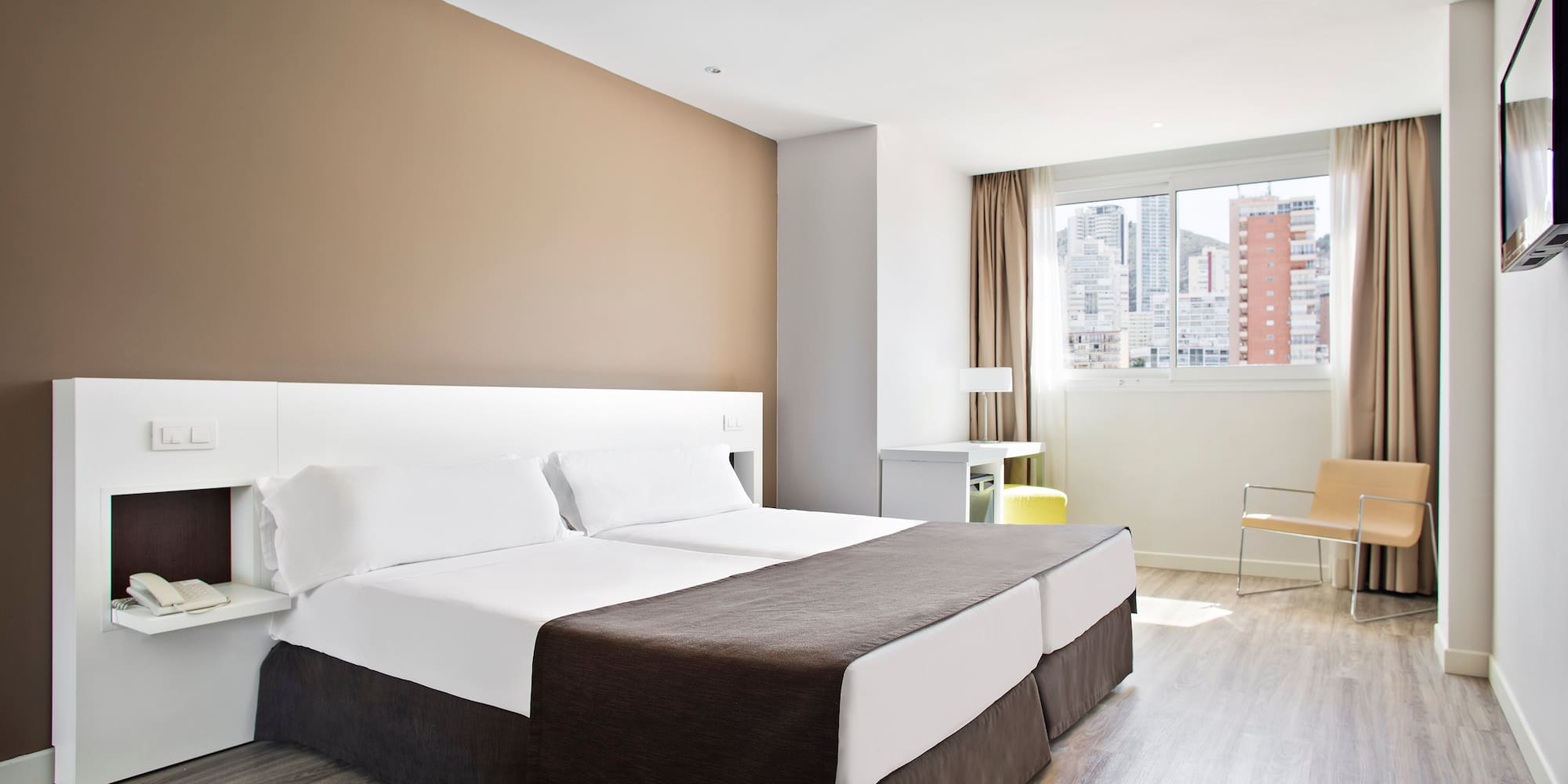 Modern hotel room with twin beds, city view, light wood floor, and bright decor.