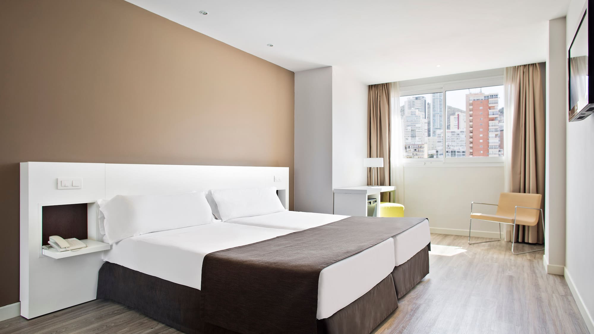 Modern hotel room with twin beds, city view, light wood floor, and bright decor.