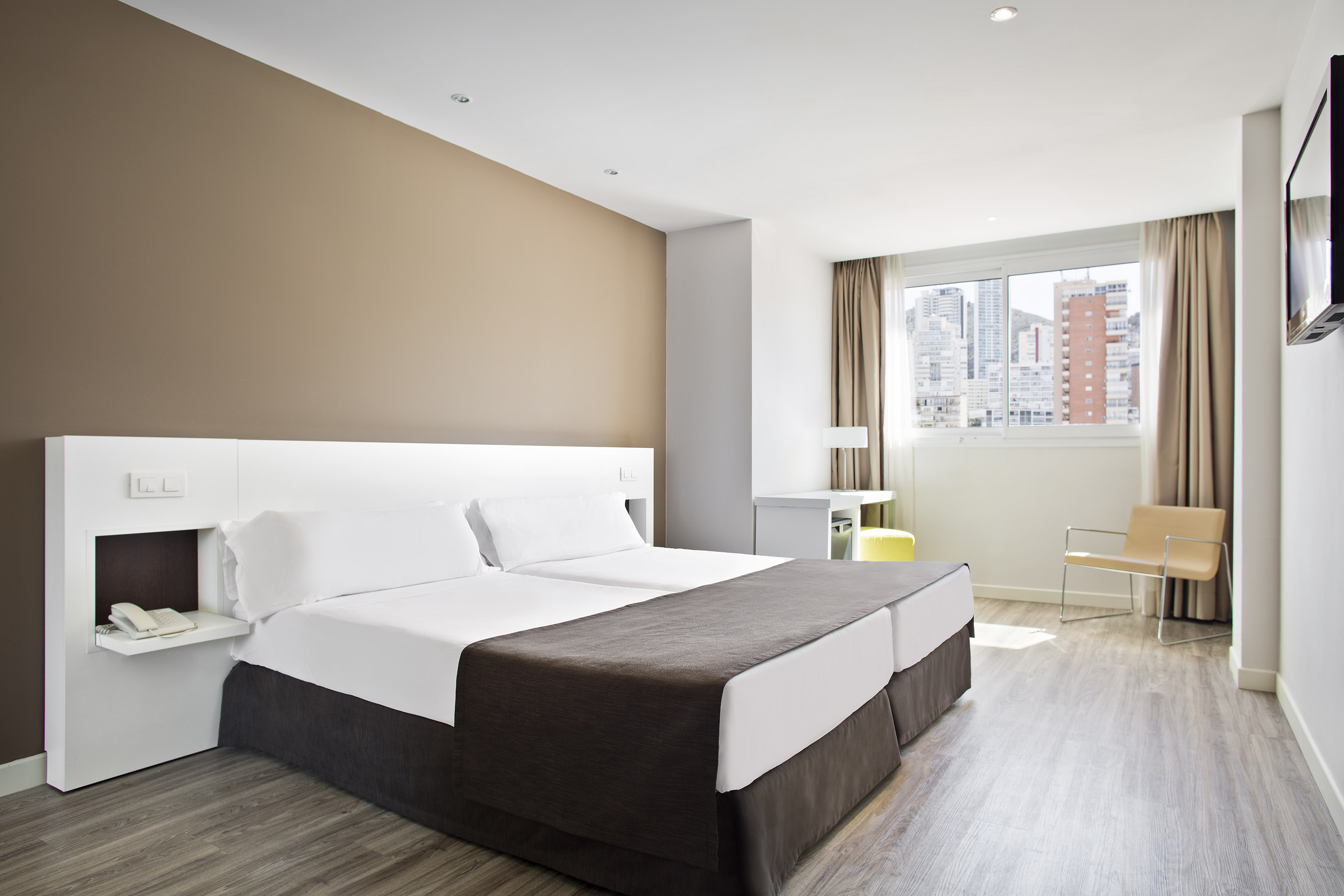 Modern hotel room with twin beds, city view, light wood floor, and bright decor.