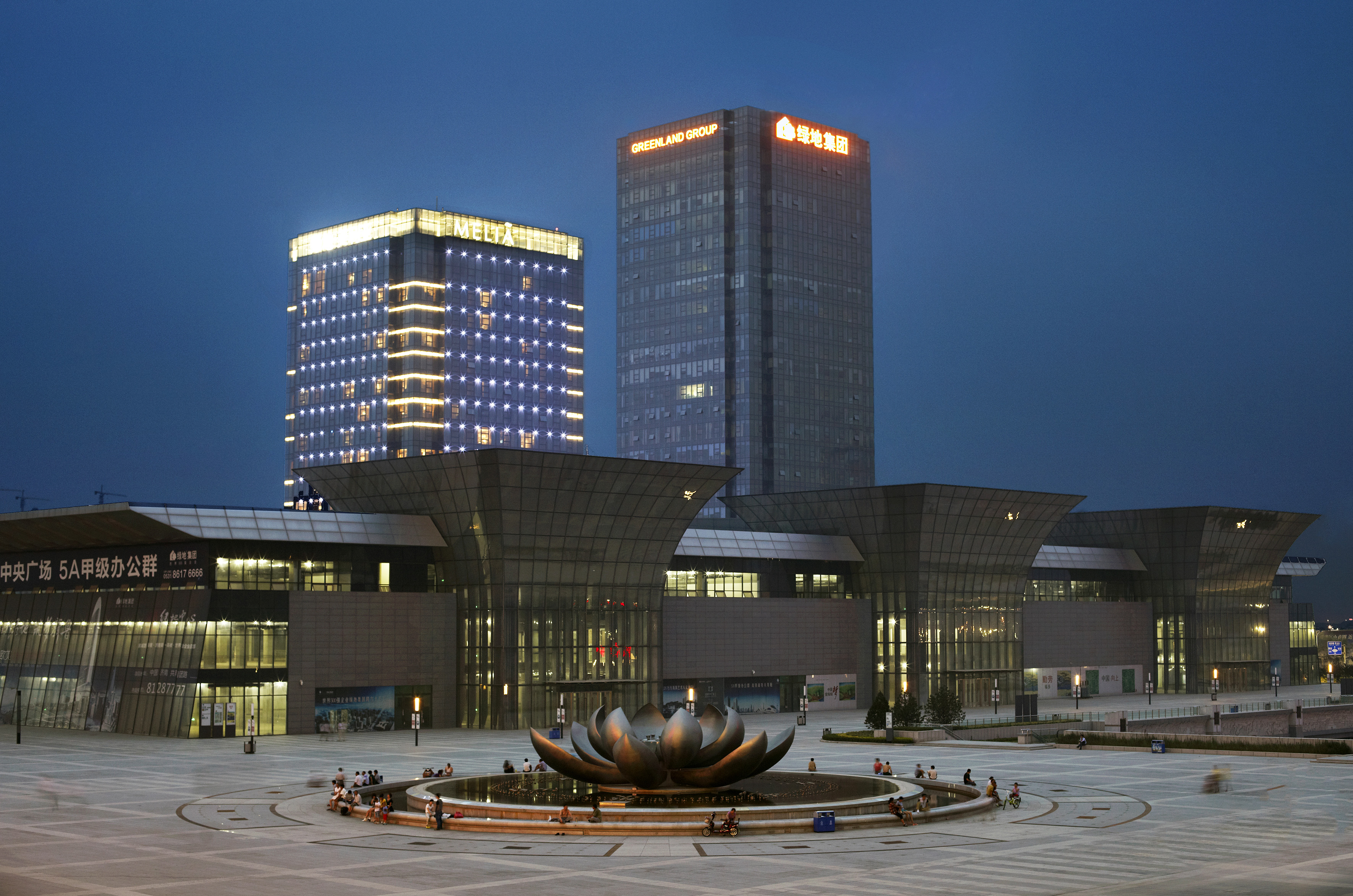 Modern city square at night with illuminated Greenland Group buildings and lotus fountain.
