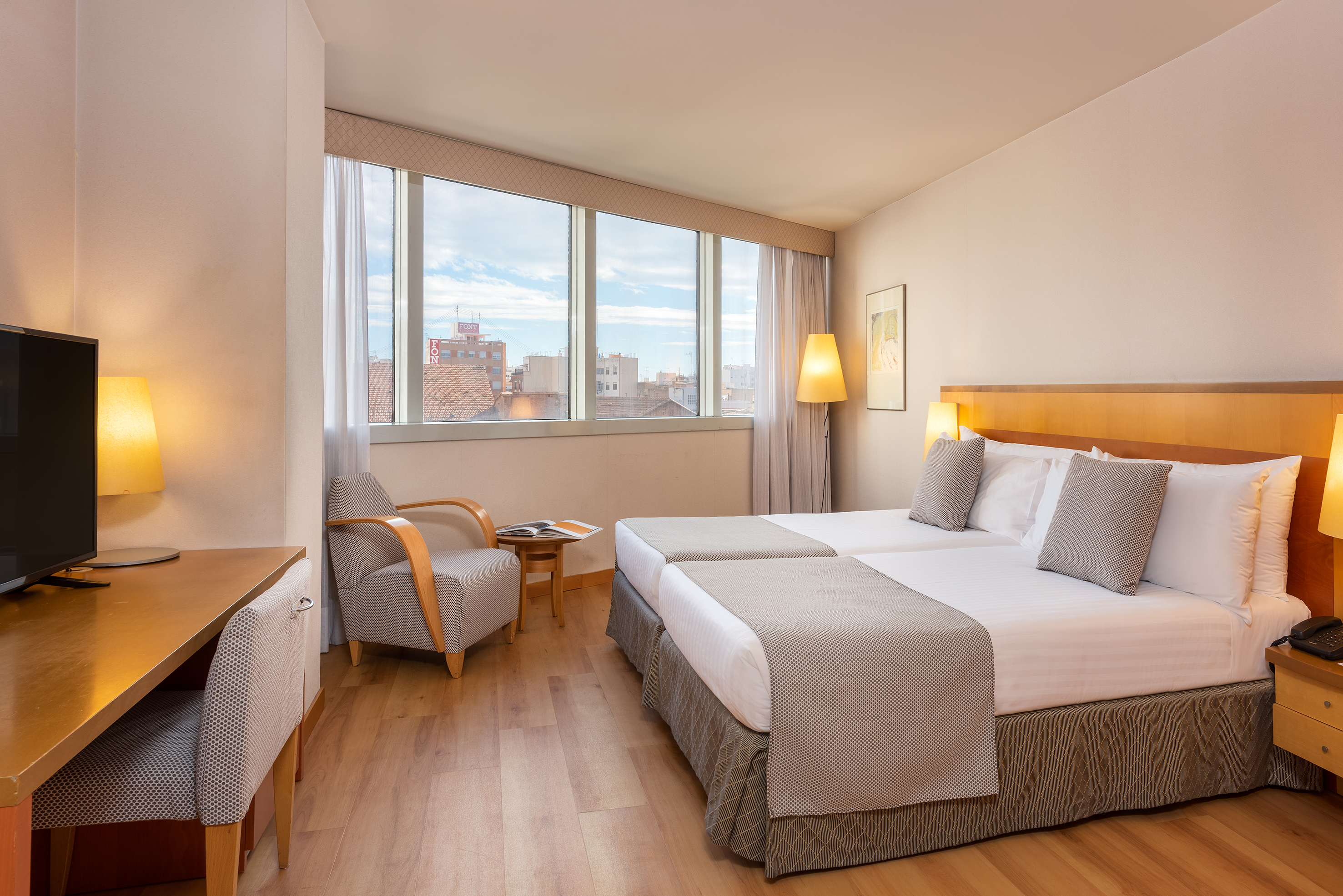 Bright hotel room with twin beds, desk, armchair, and city view.