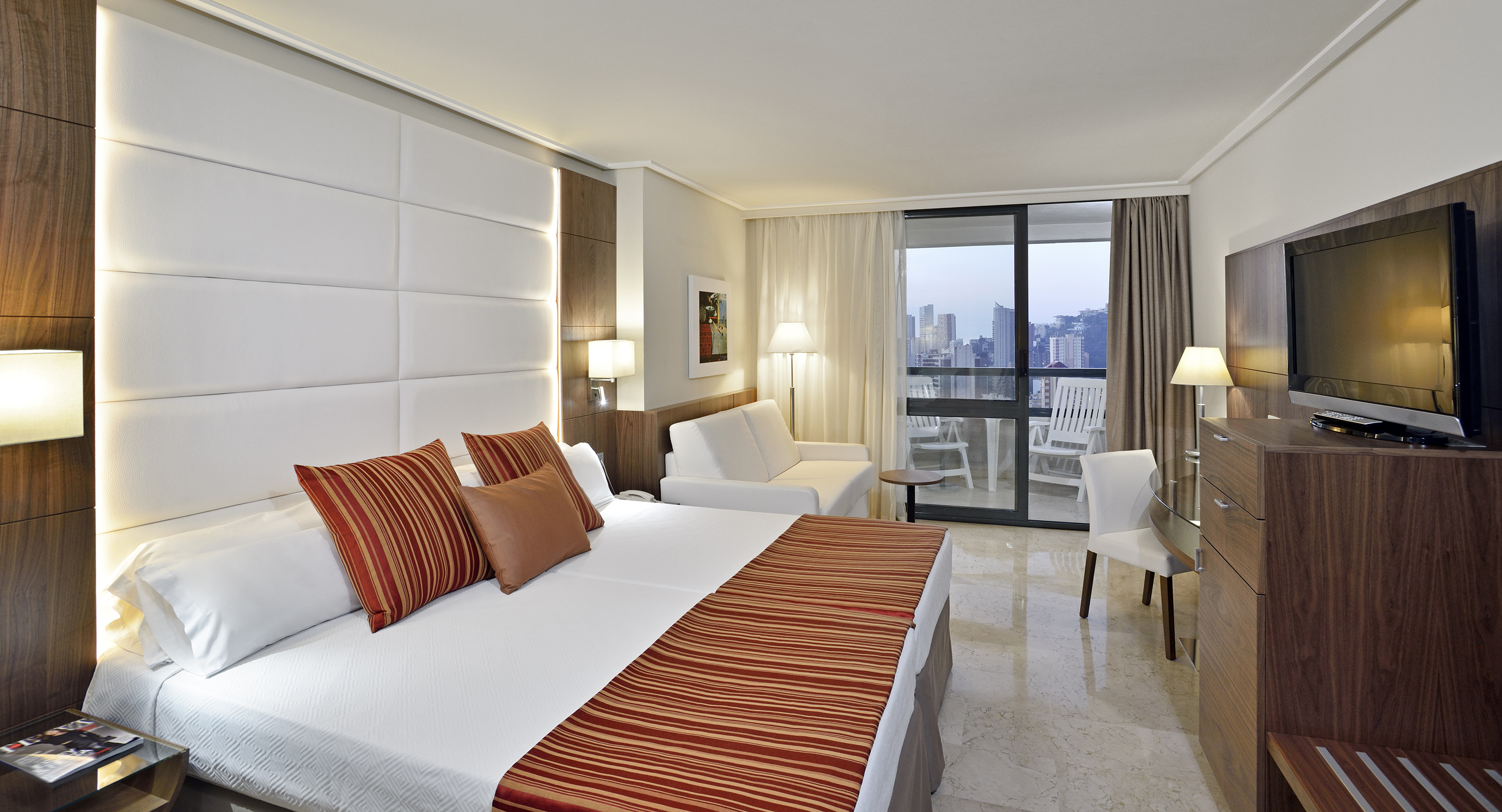 Hotel room with king bed, white headboard, city view balcony, and sofa.