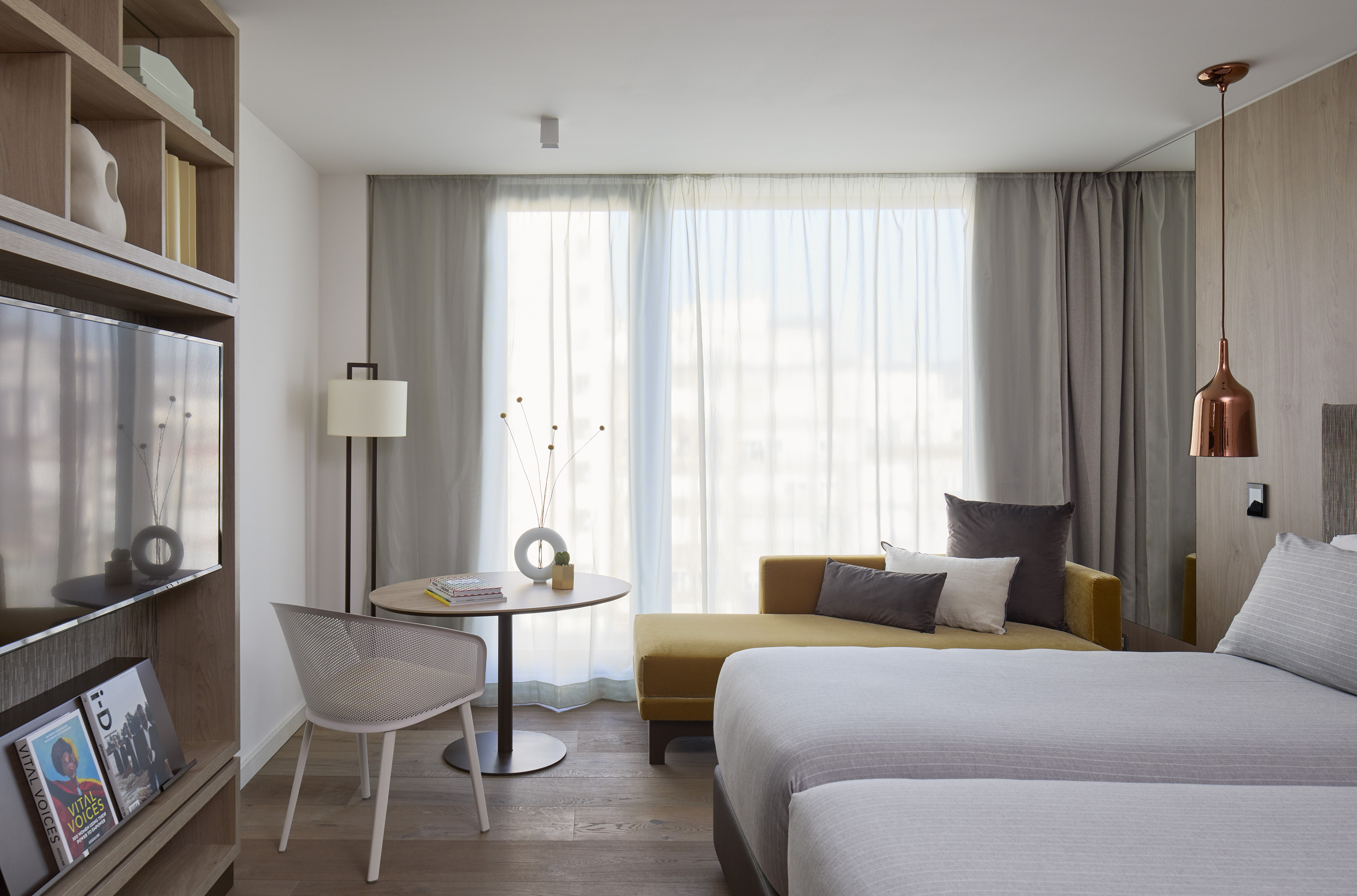Modern hotel room with bed, yellow lounge, wooden floor, and sheer curtains.