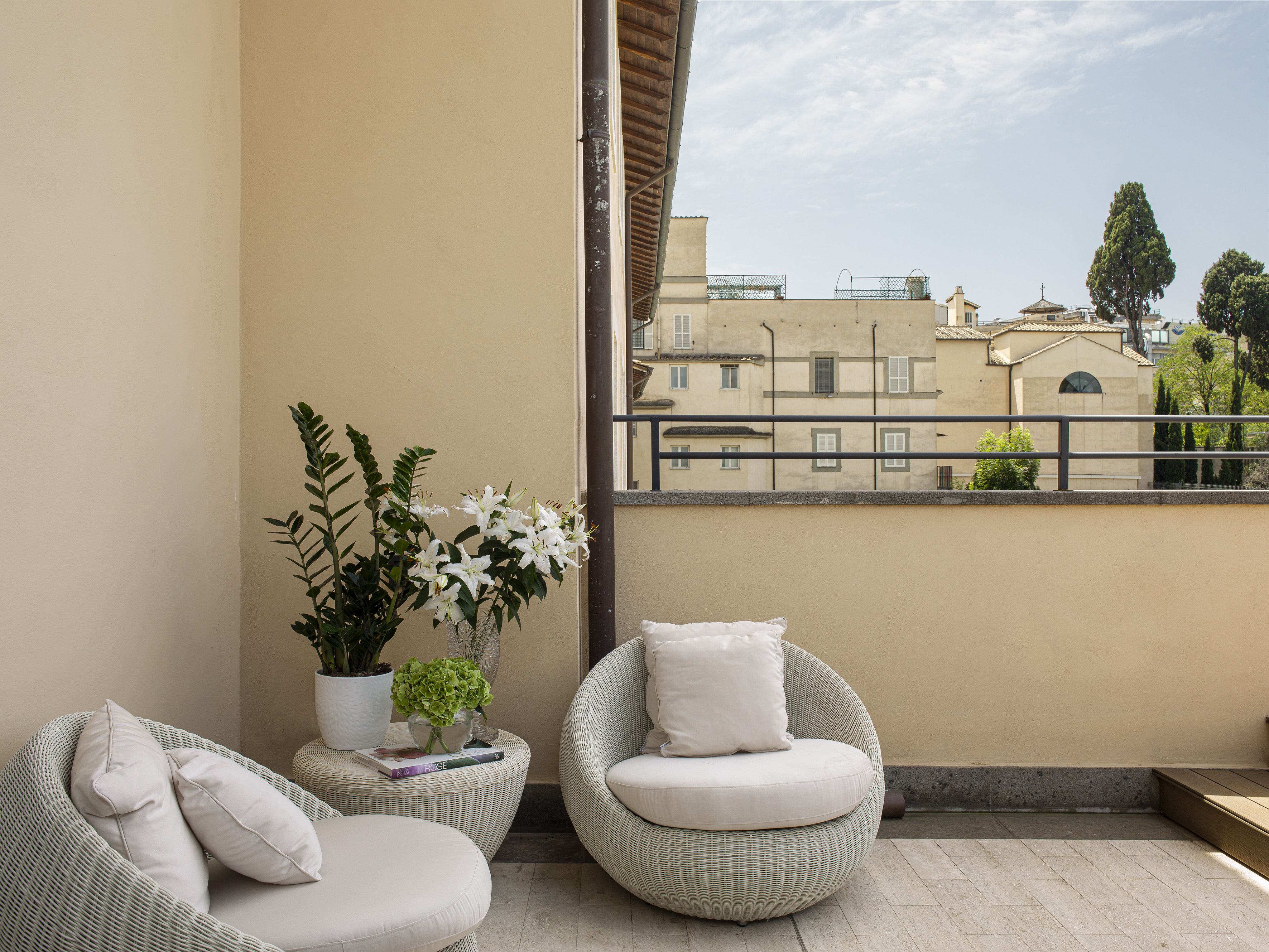 Balcony terrace with wicker seating, potted plants, and distant city buildings.