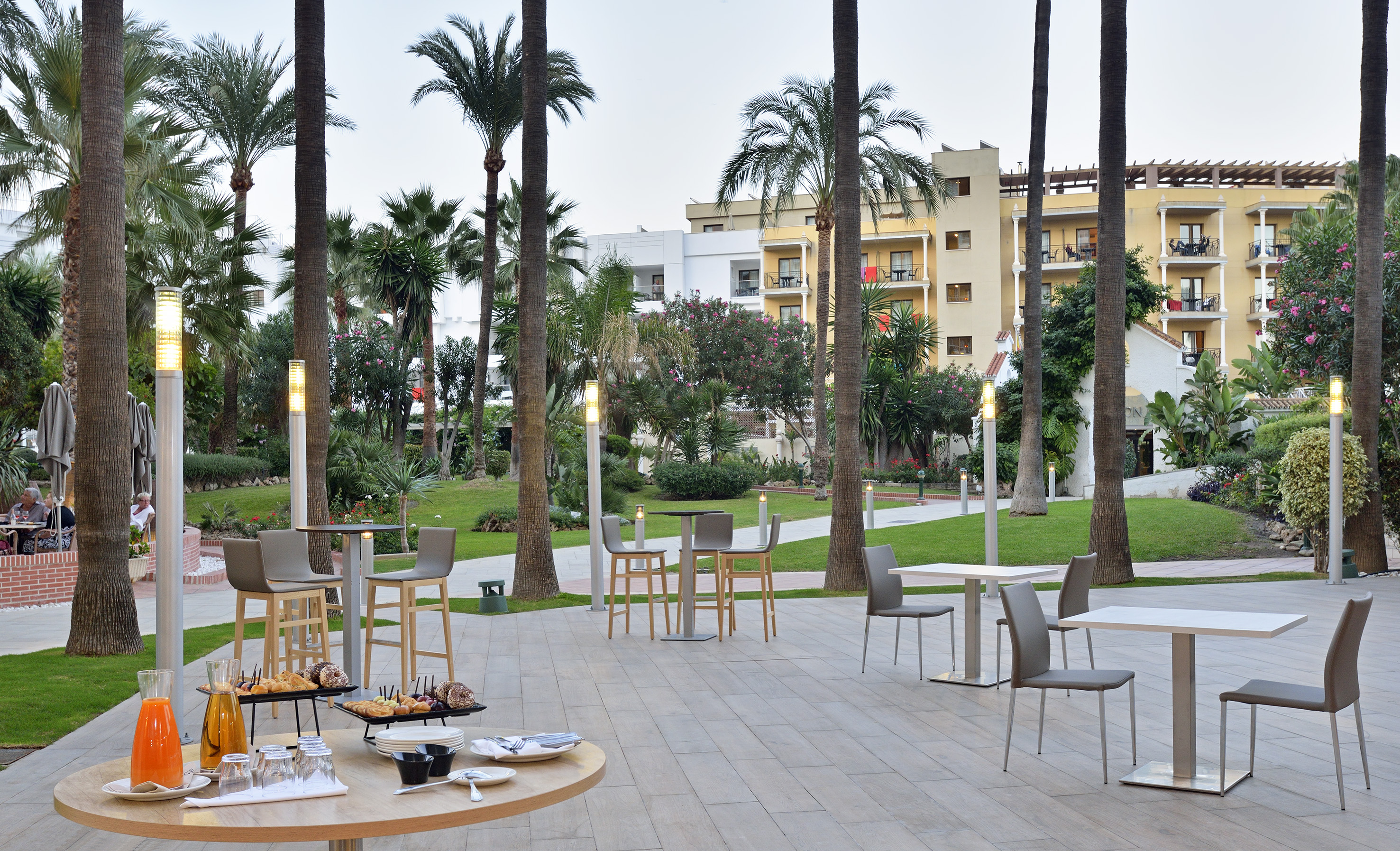 Outdoor resort terrace with palm trees, breakfast display, and hotel backdrop.