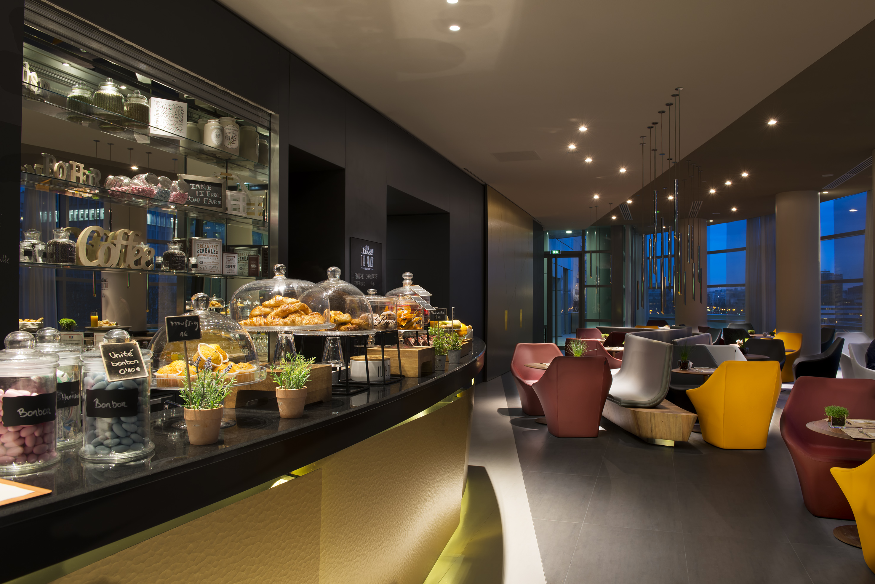 Modern cafe bar with pastries, coffee, colorful lounge, and evening city view.