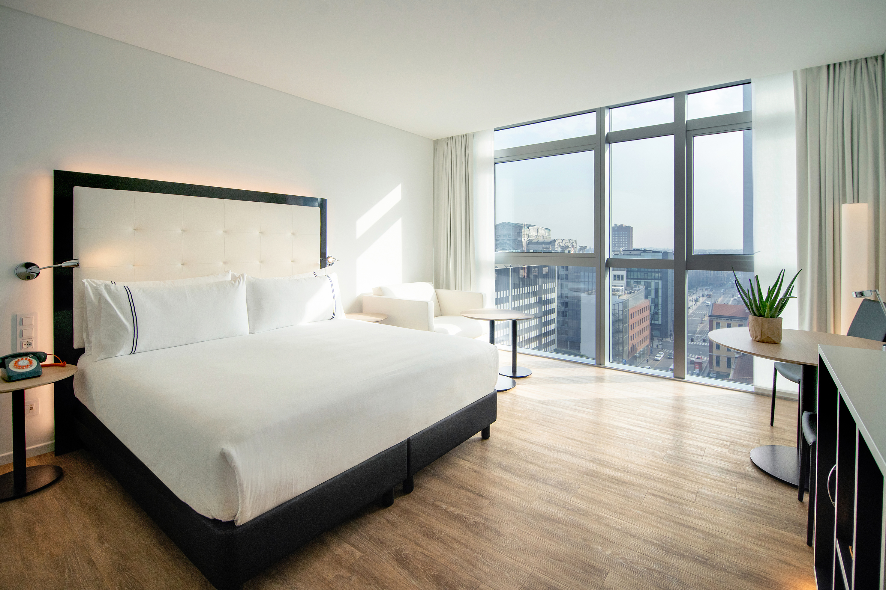Bright modern hotel bedroom with large bed, city view, and wooden floor.