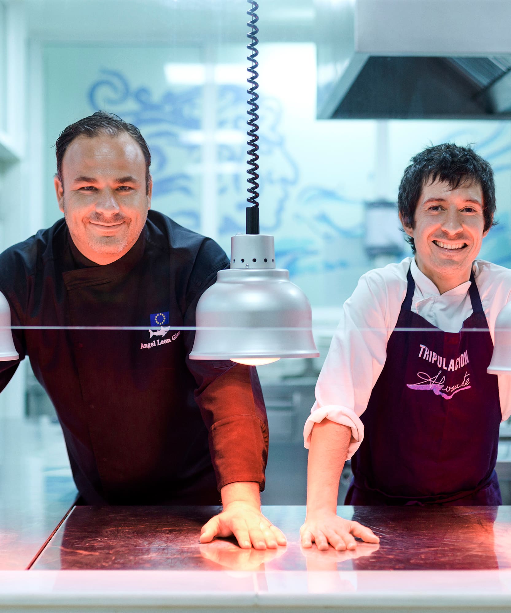 two men in aprons standing behind a counter with lights