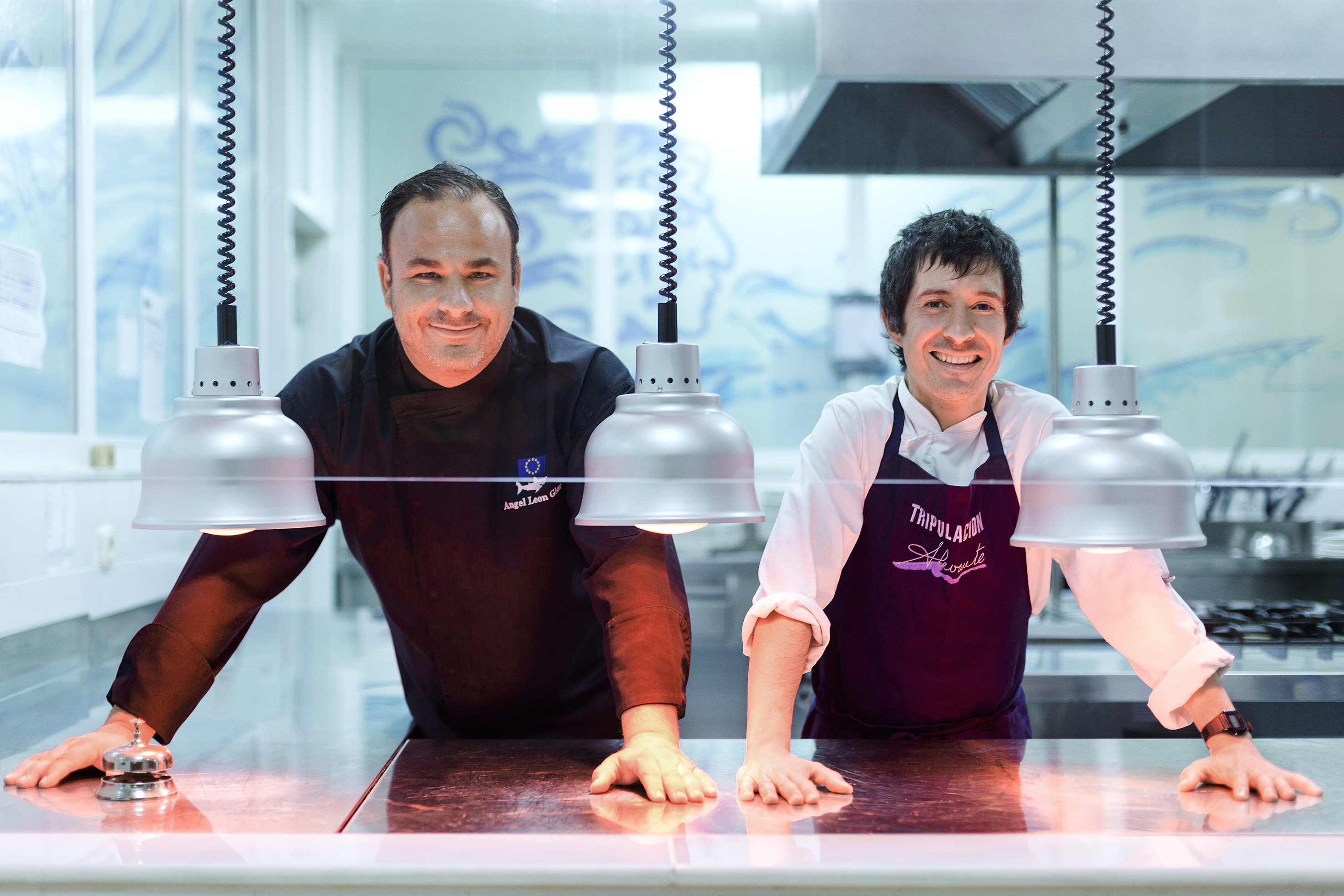 two men in aprons standing behind a counter with lights