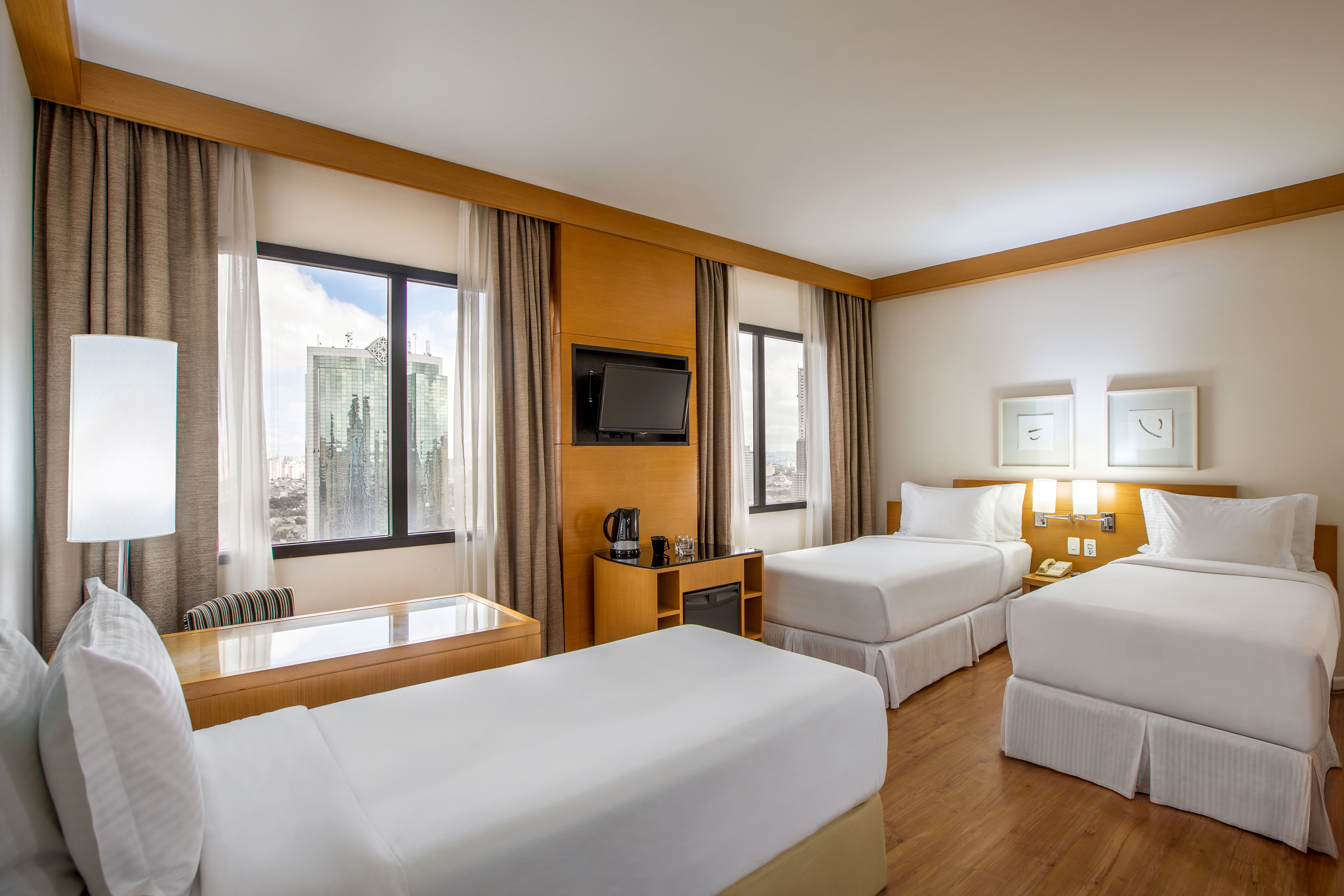 Bright hotel room with three twin beds, city view windows, TV, and work desk.