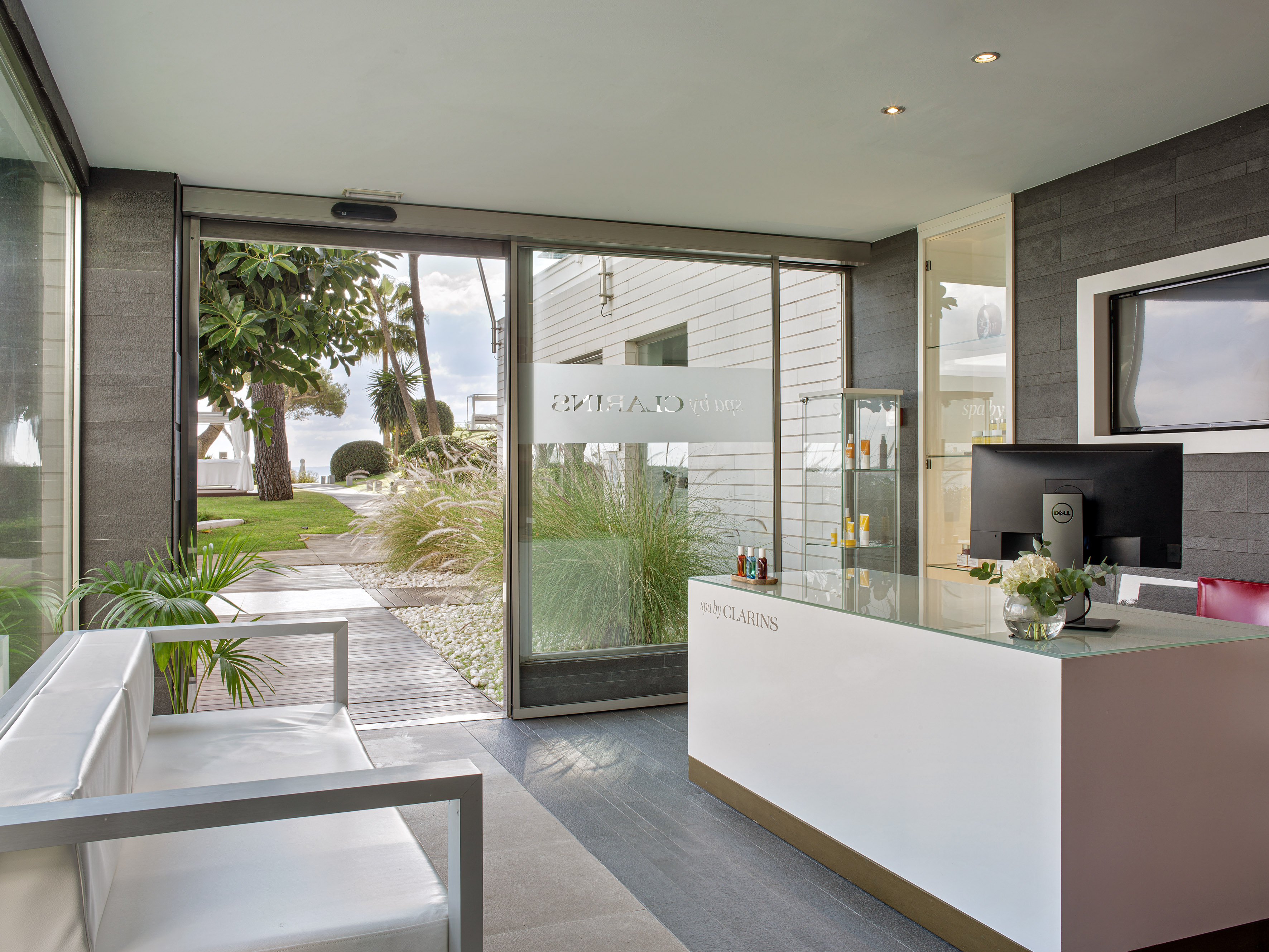 Modern Clarins spa reception with white desk, sofa, and outdoor garden view.