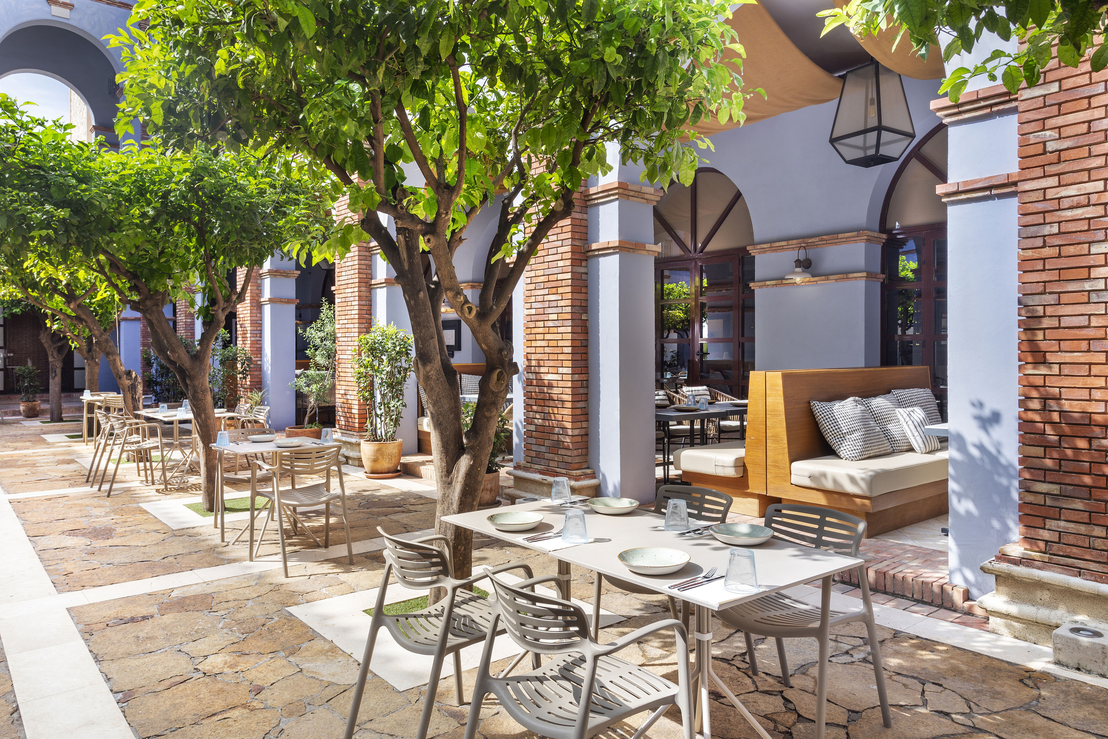 Shaded restaurant courtyard with dining tables, leafy trees, and brick walls.