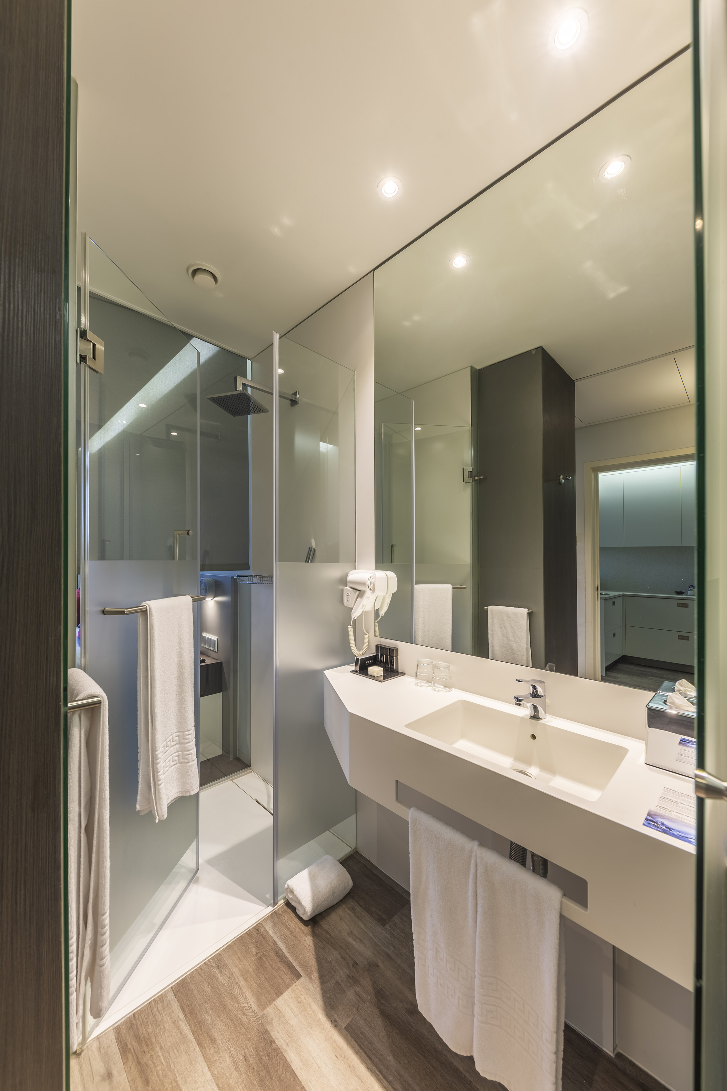 Sleek modern bathroom with glass shower, white vanity, large mirror, and wood floor. - Classic Room - Meliá Maputo Sky, Maputo