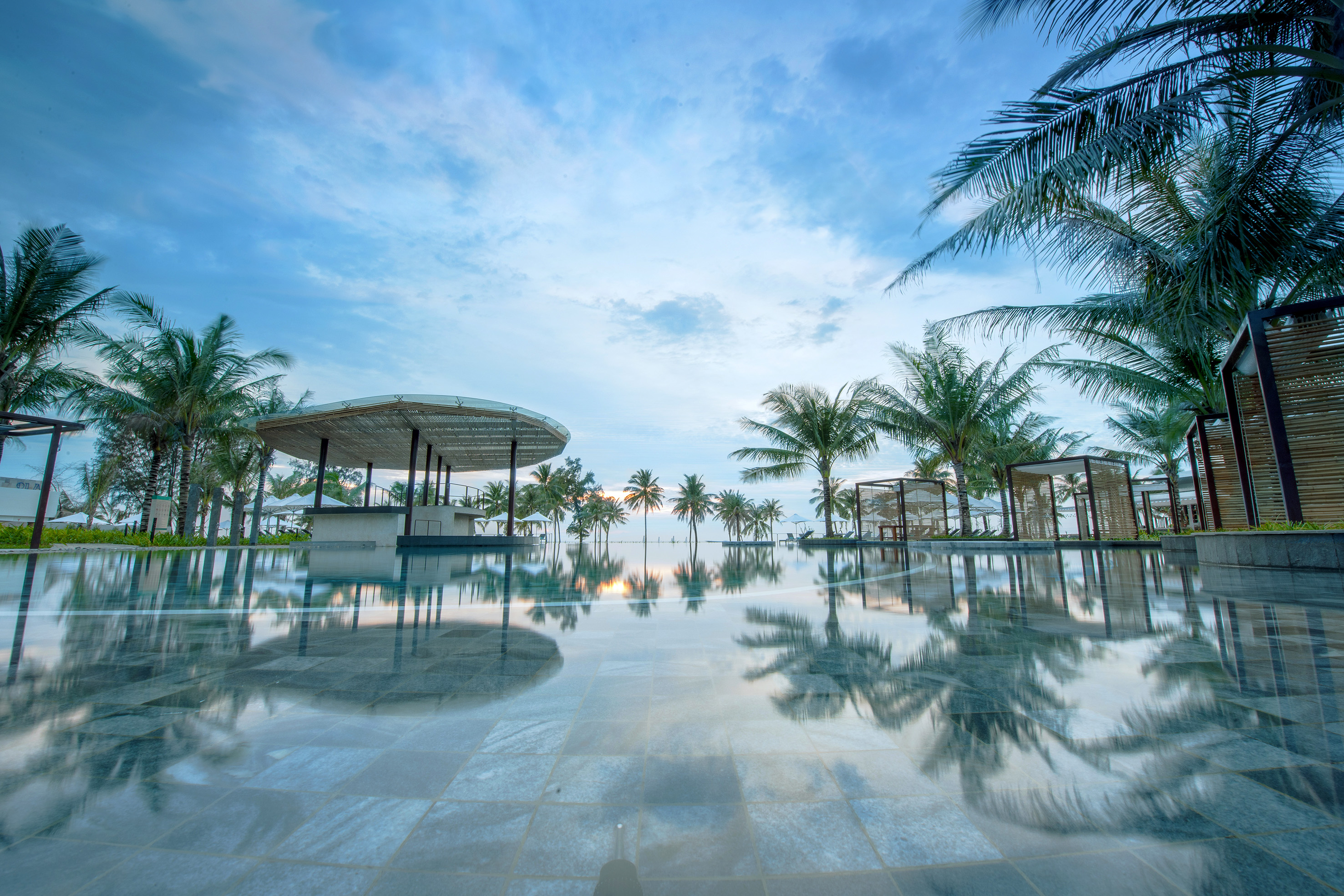 Tropical resort pool reflecting palm trees and outdoor bar at sunrise.