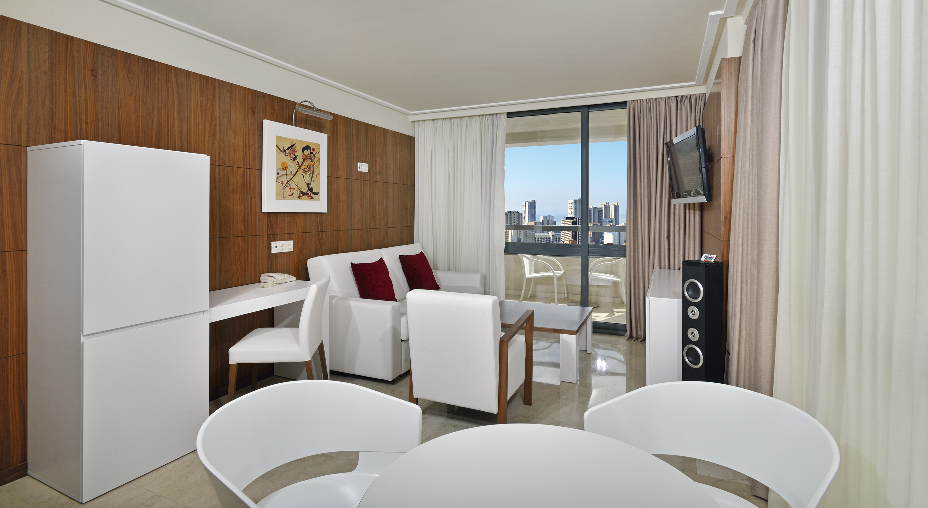 Modern hotel suite with white furniture, wooden walls, city view balcony.