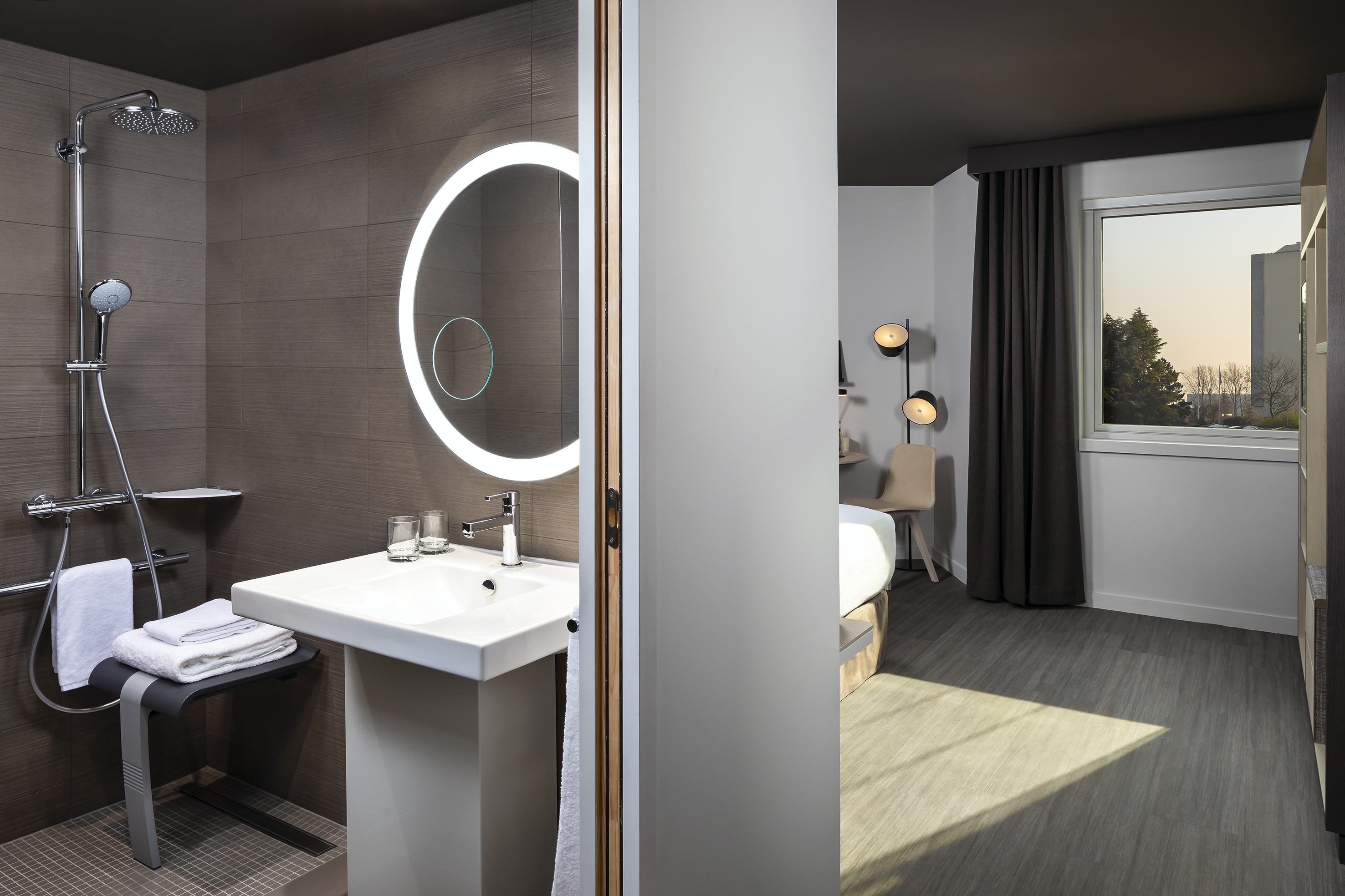Modern hotel room: accessible shower, lighted mirror, vanity, and bedroom.