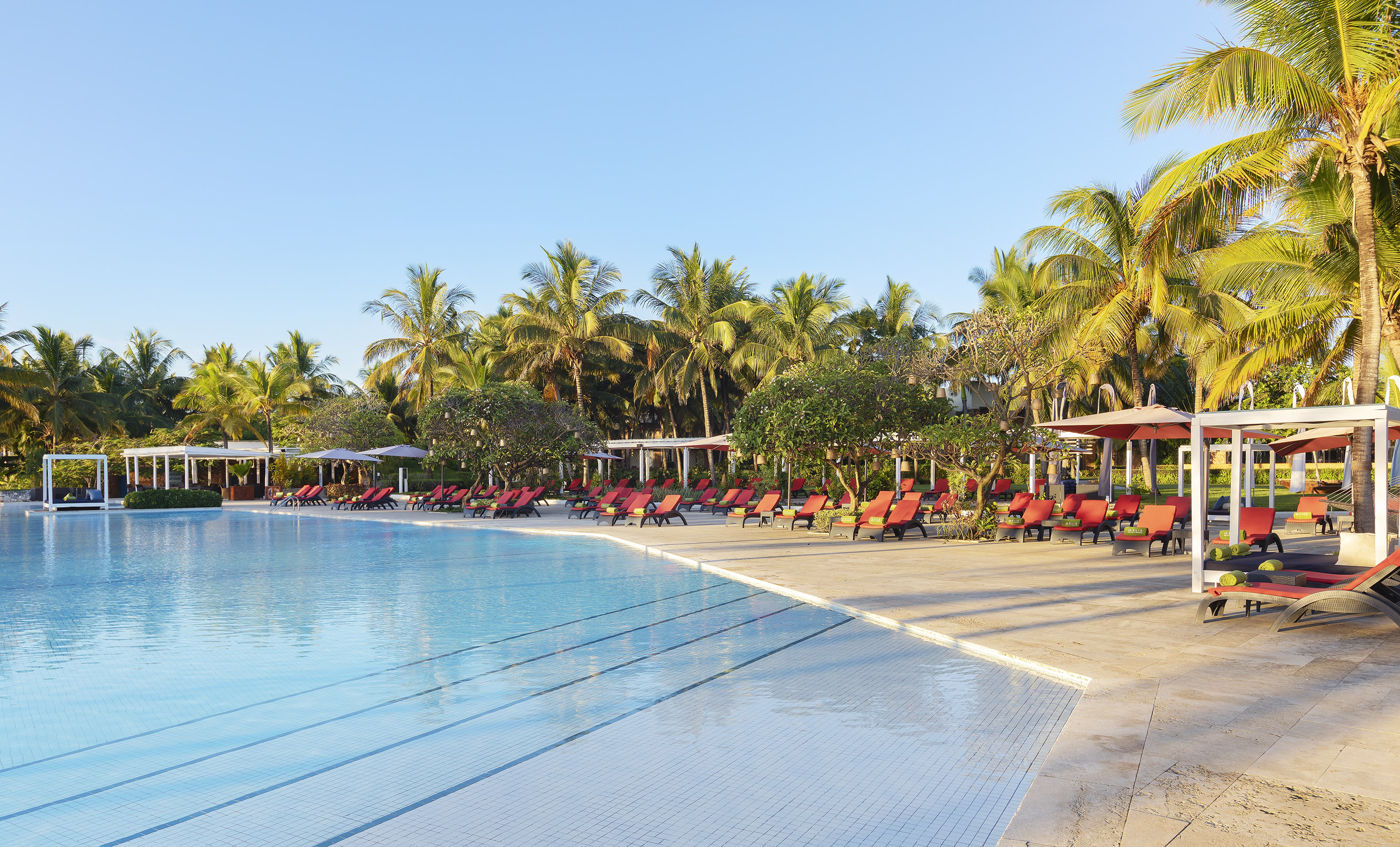 Tropical resort pool with red sun loungers, cabanas, and palm trees.