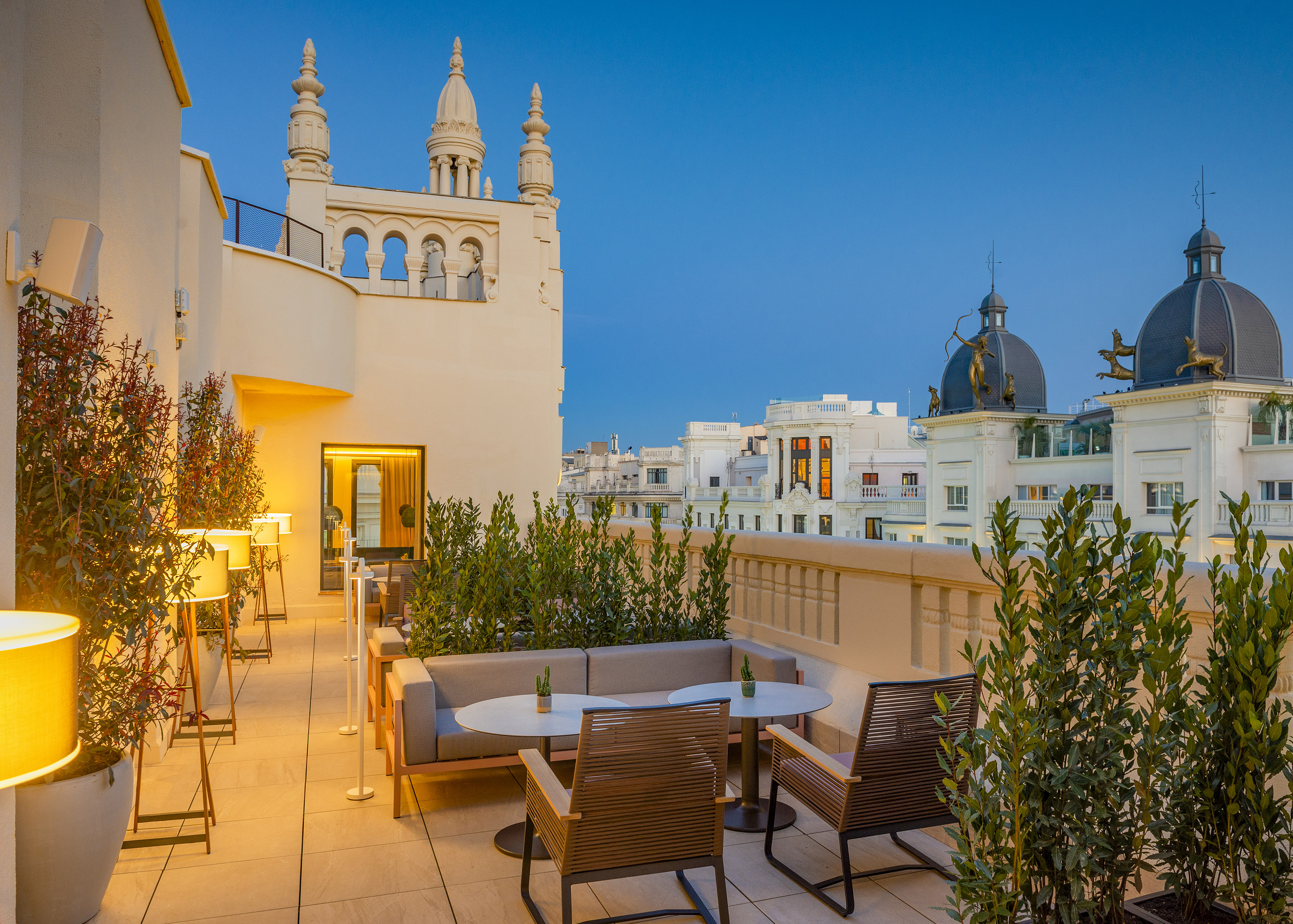Rooftop terrace with outdoor lounge seating, lush plants, and evening city view.