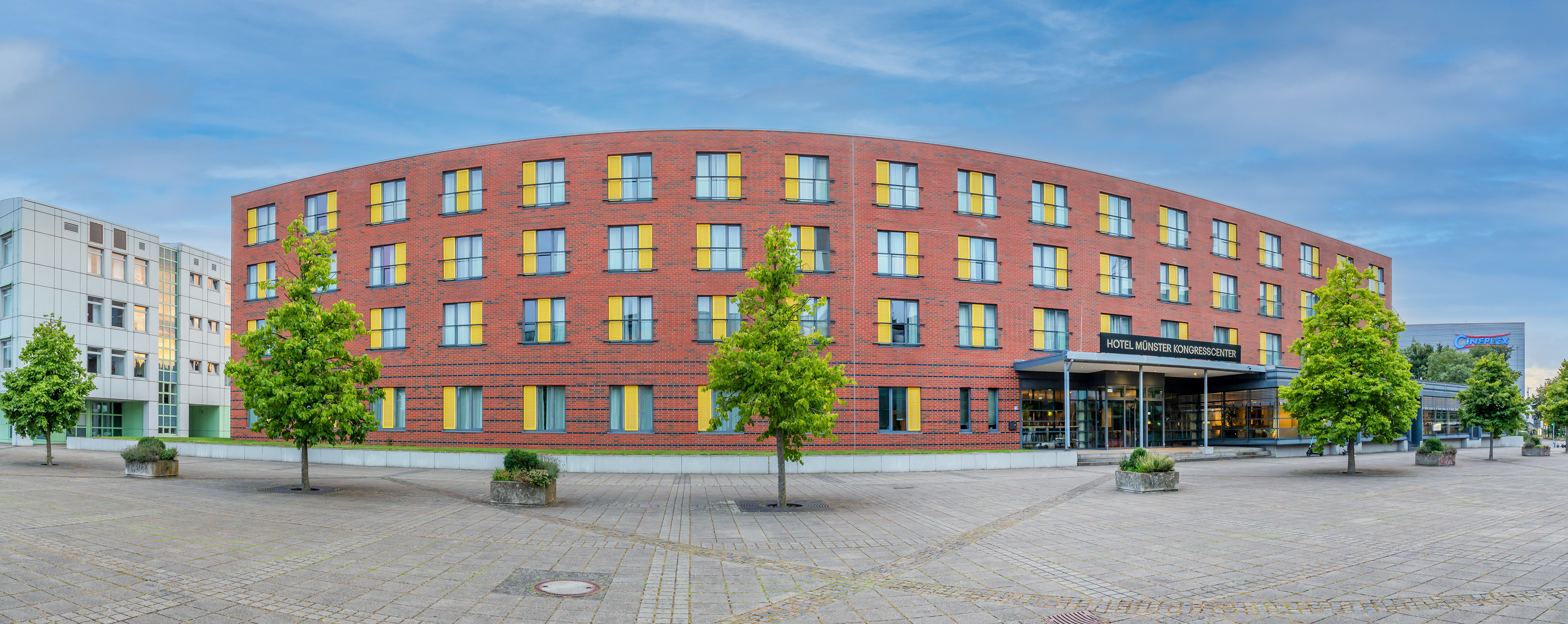 Curved brick Hotel Münster Kongresscenter with yellow windows on a paved plaza.