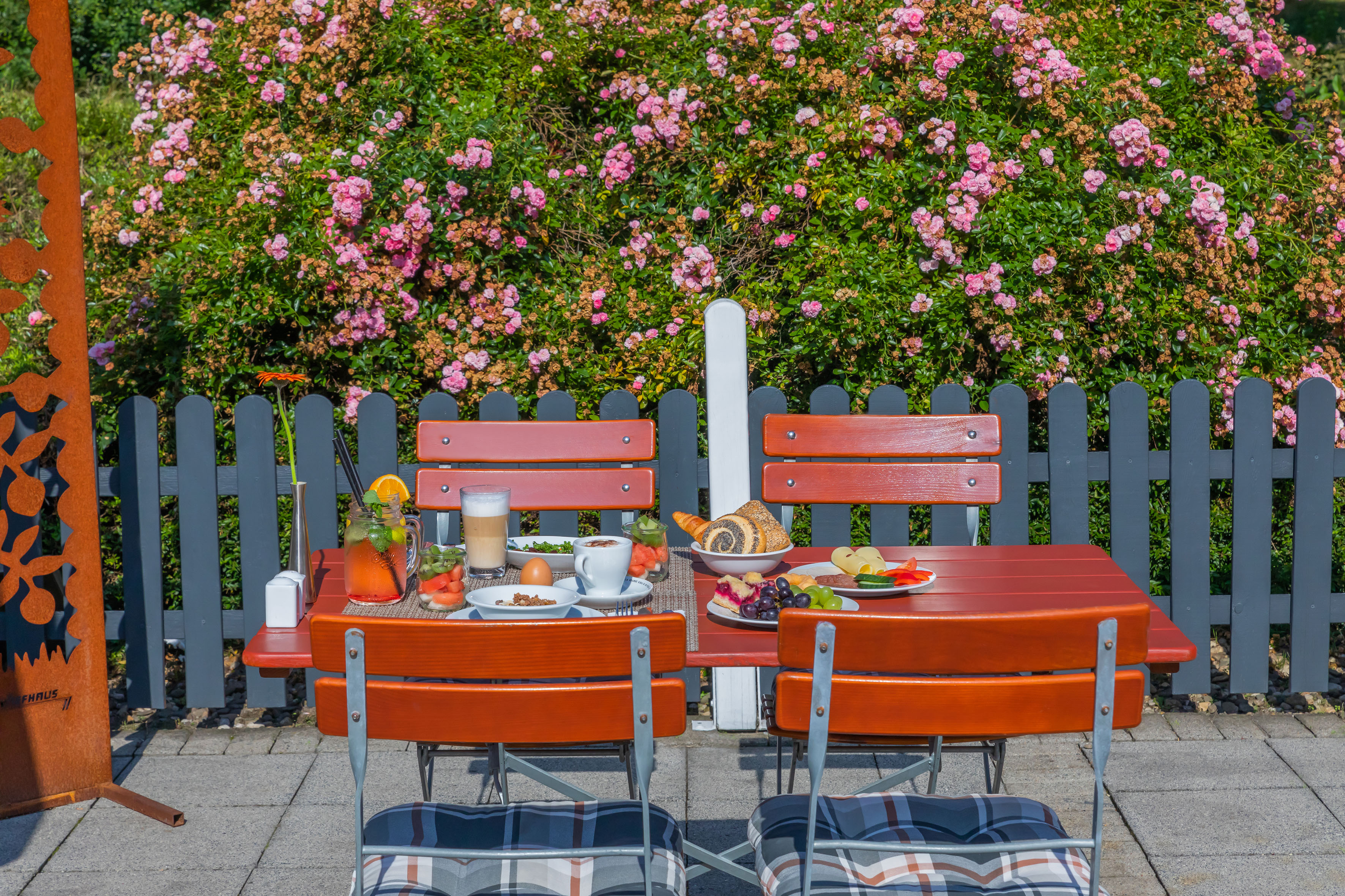 Sunny outdoor brunch with coffee, pastries, fruit, and blooming pink roses.
